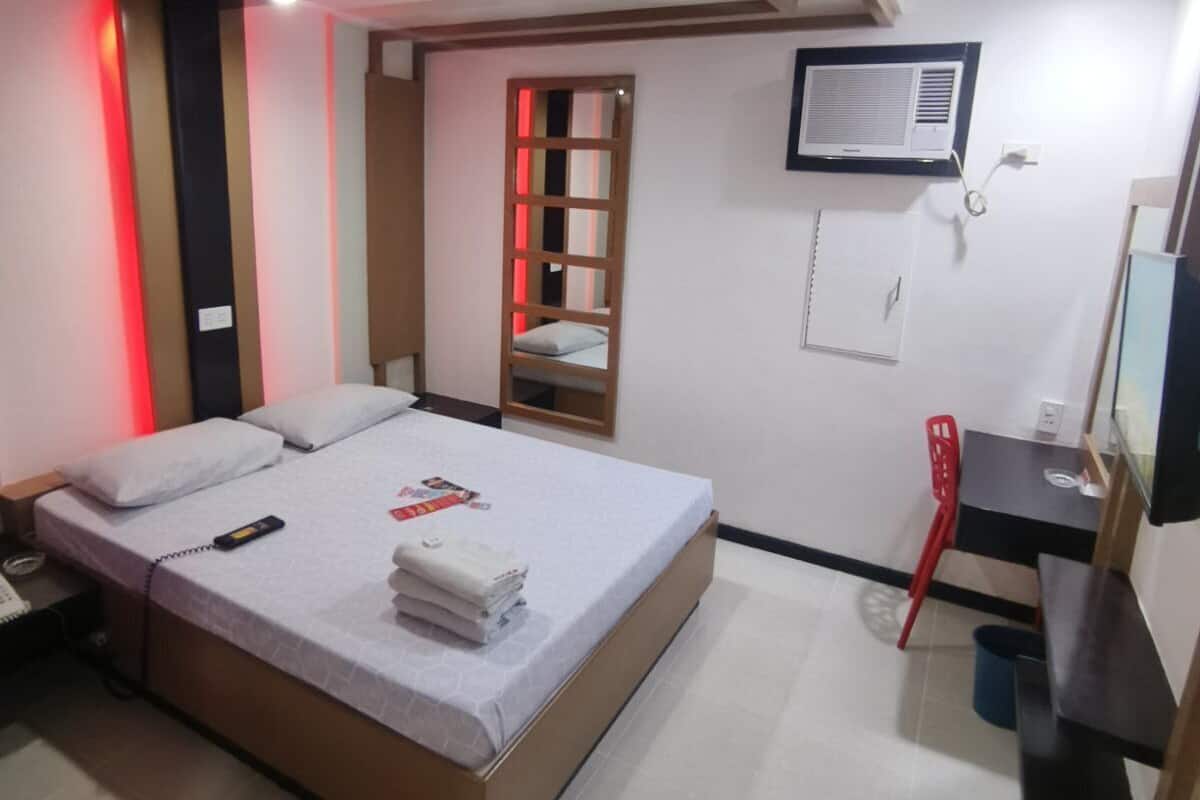 A hotel room with a bed, a desk, a chair, an air conditioning unit, and a television.