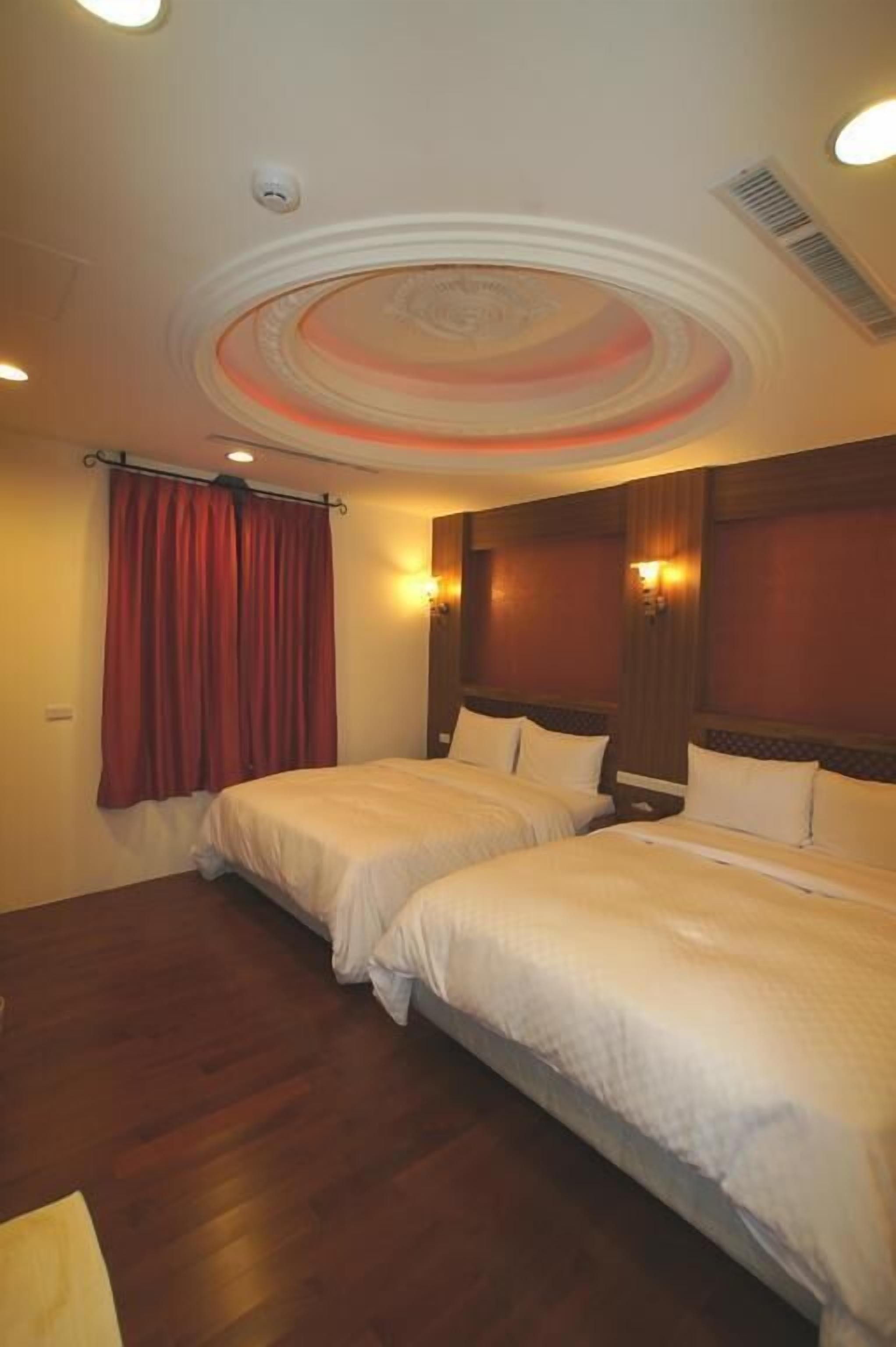 Una habitación de hotel con dos camas, suelo de madera, cortina roja y techo decorativo.
