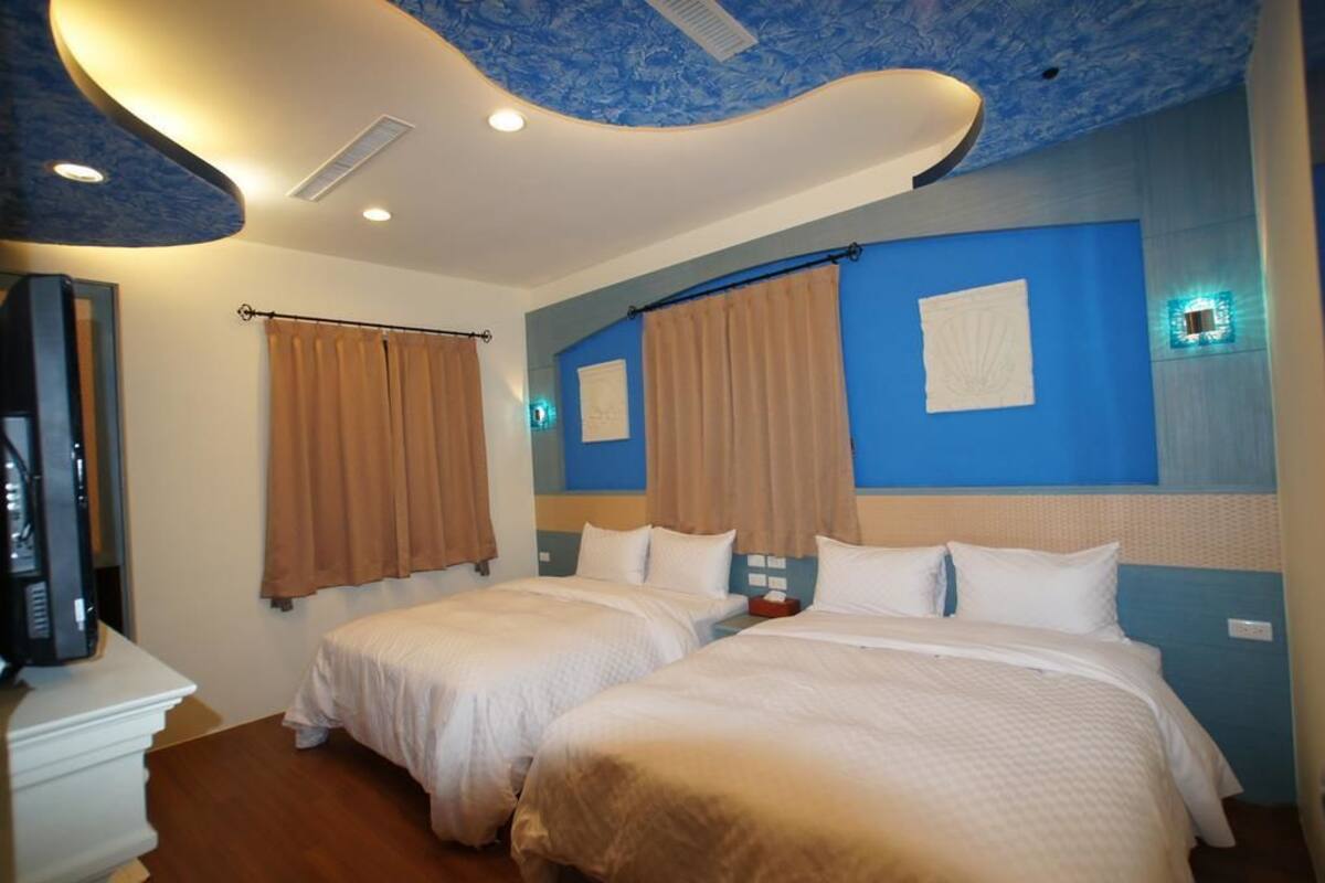 A hotel room with two beds, beige curtains, a flat-screen TV, and a ceiling with a blue pattern.