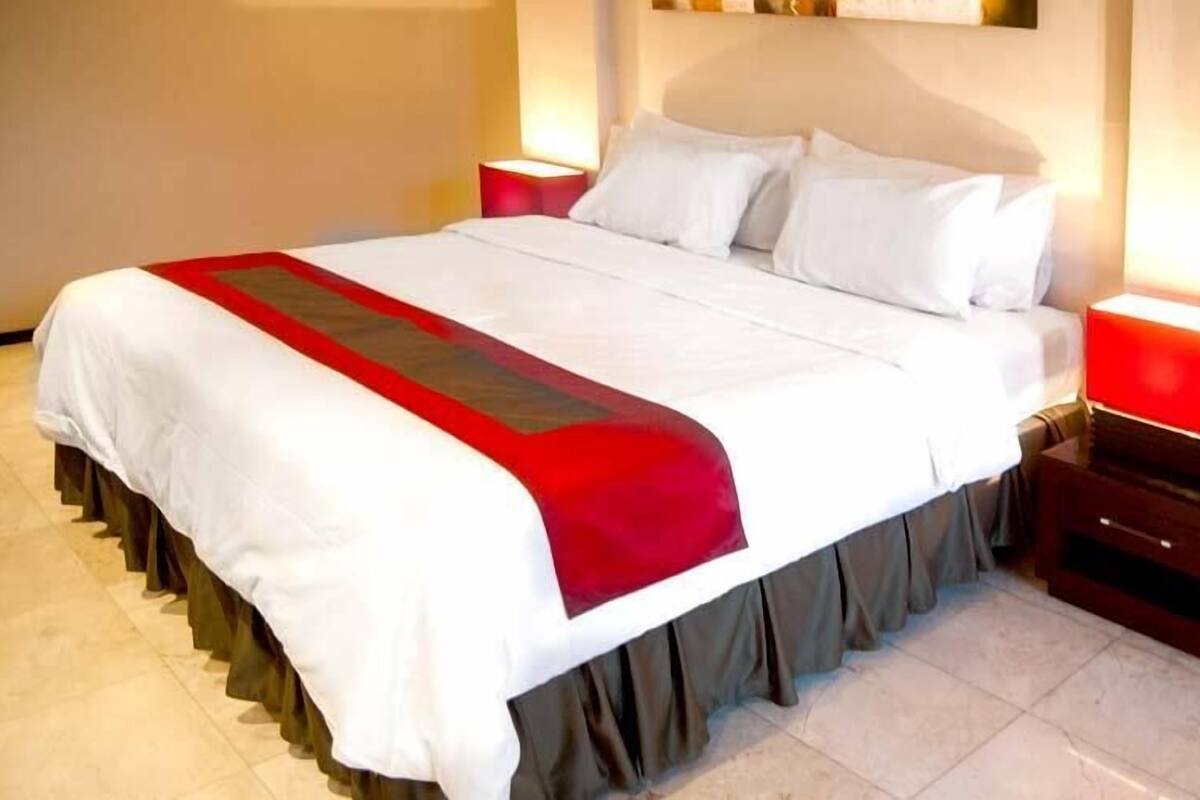 A neatly made bed with white bedding and a red accent, two bedside tables with red lamps, and a modern headboard.