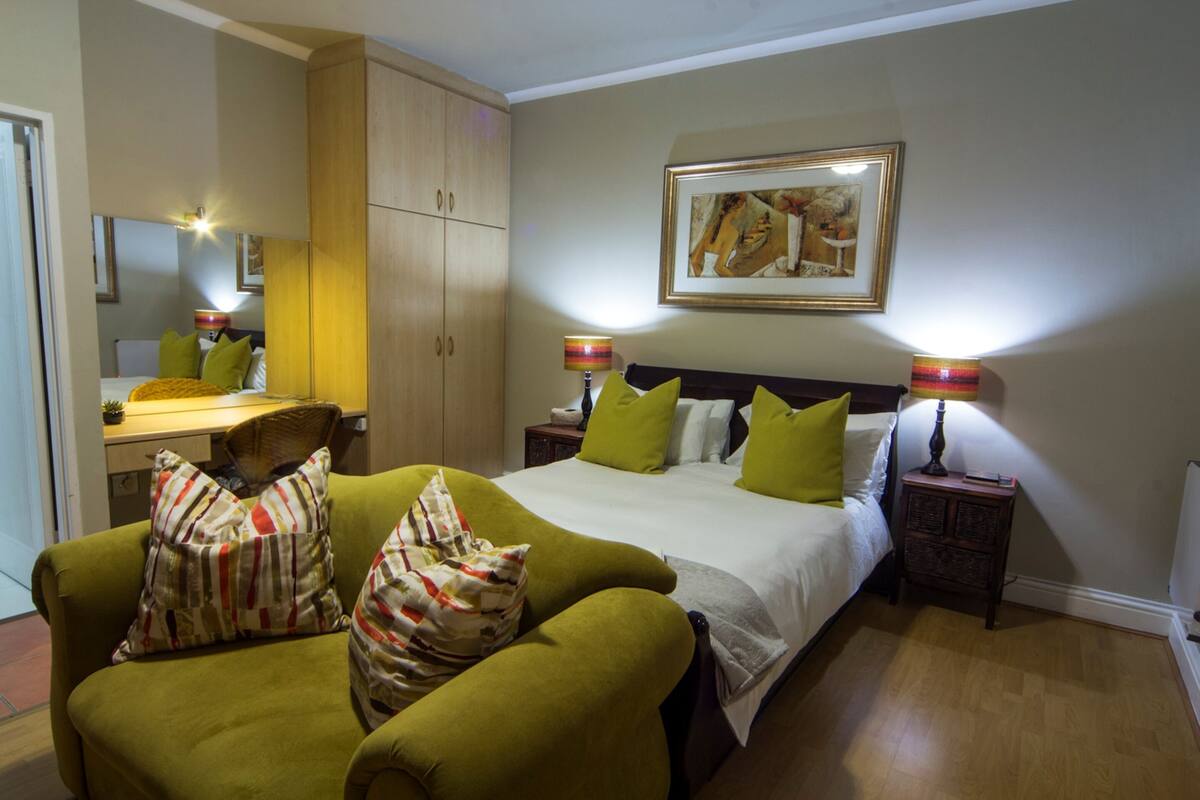 Een hotelkamer met een bed, groene fauteuils, een houten garderobe, een bureau en een schilderij aan de muur.
