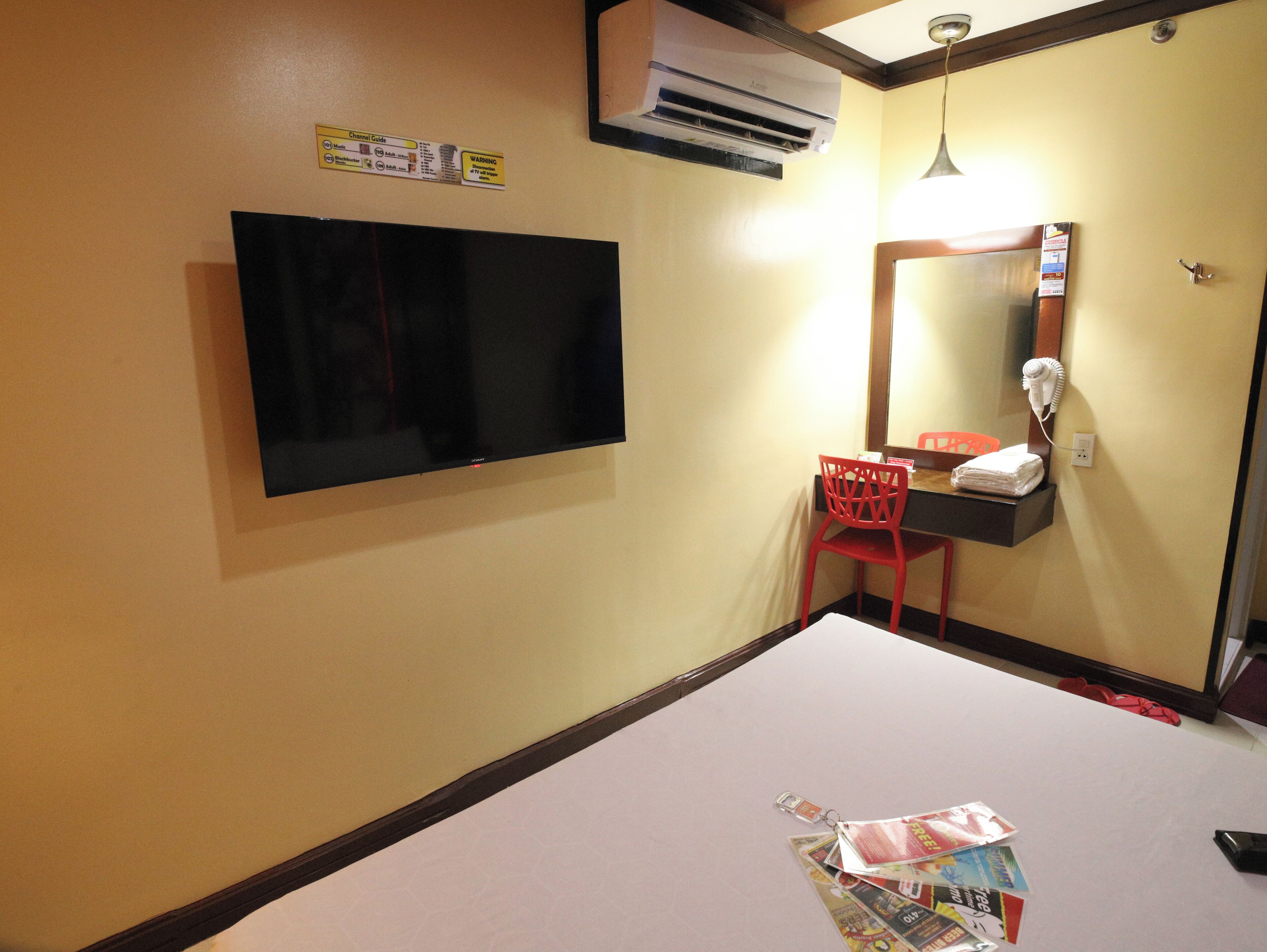 A hotel room with a flat-screen TV mounted on the wall, a red chair, a mirror with a hairdryer, and a small shelf with a towel.