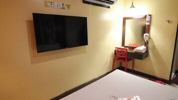 A hotel room with a flat-screen TV mounted on the wall, a red chair, a mirror with a hairdryer, and a small shelf with a towel.