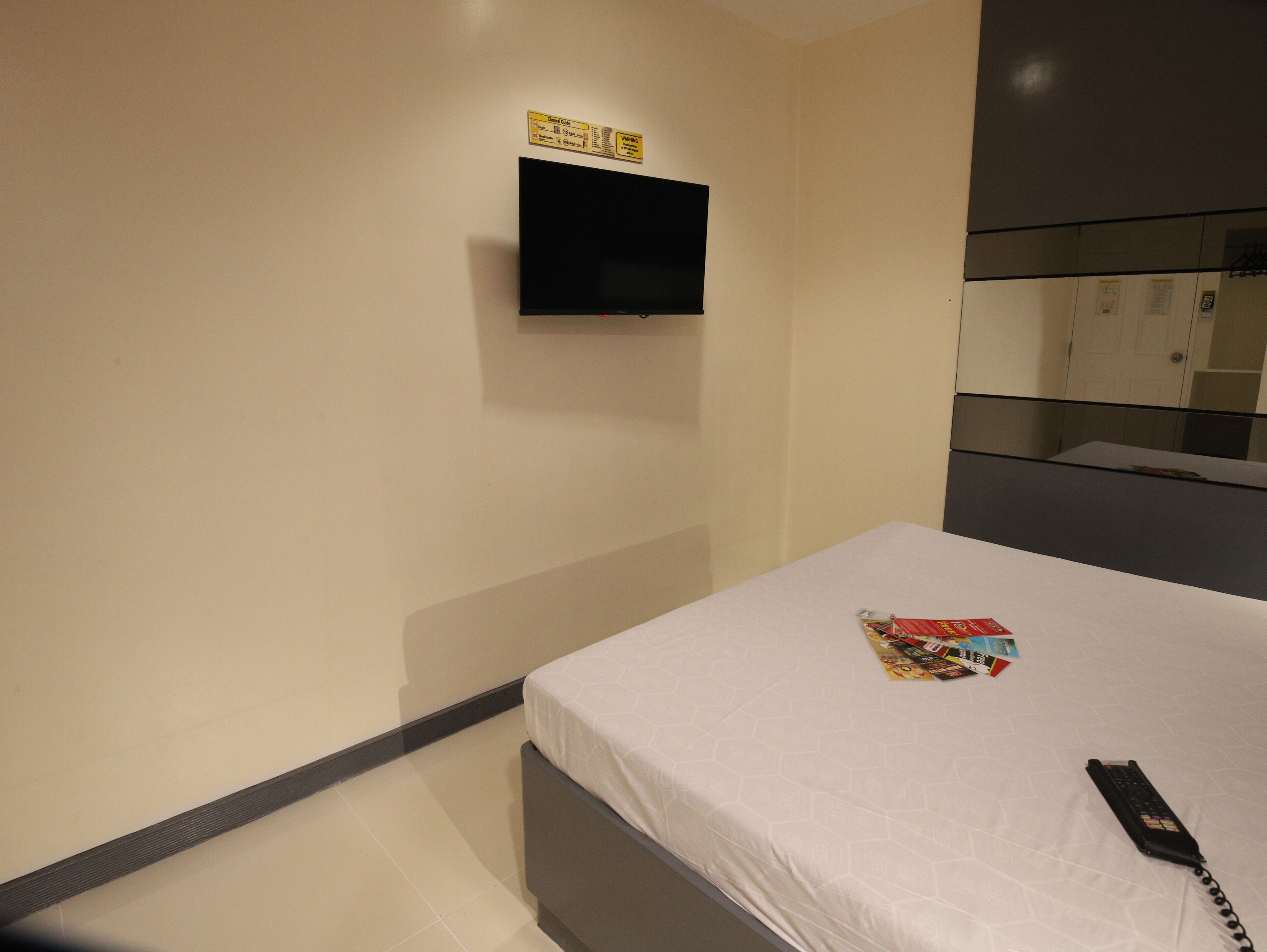 Habitación de hotel con cama, televisor de pantalla plana en la pared, teléfono sobre la cama y una pequeña mesa con una revista.