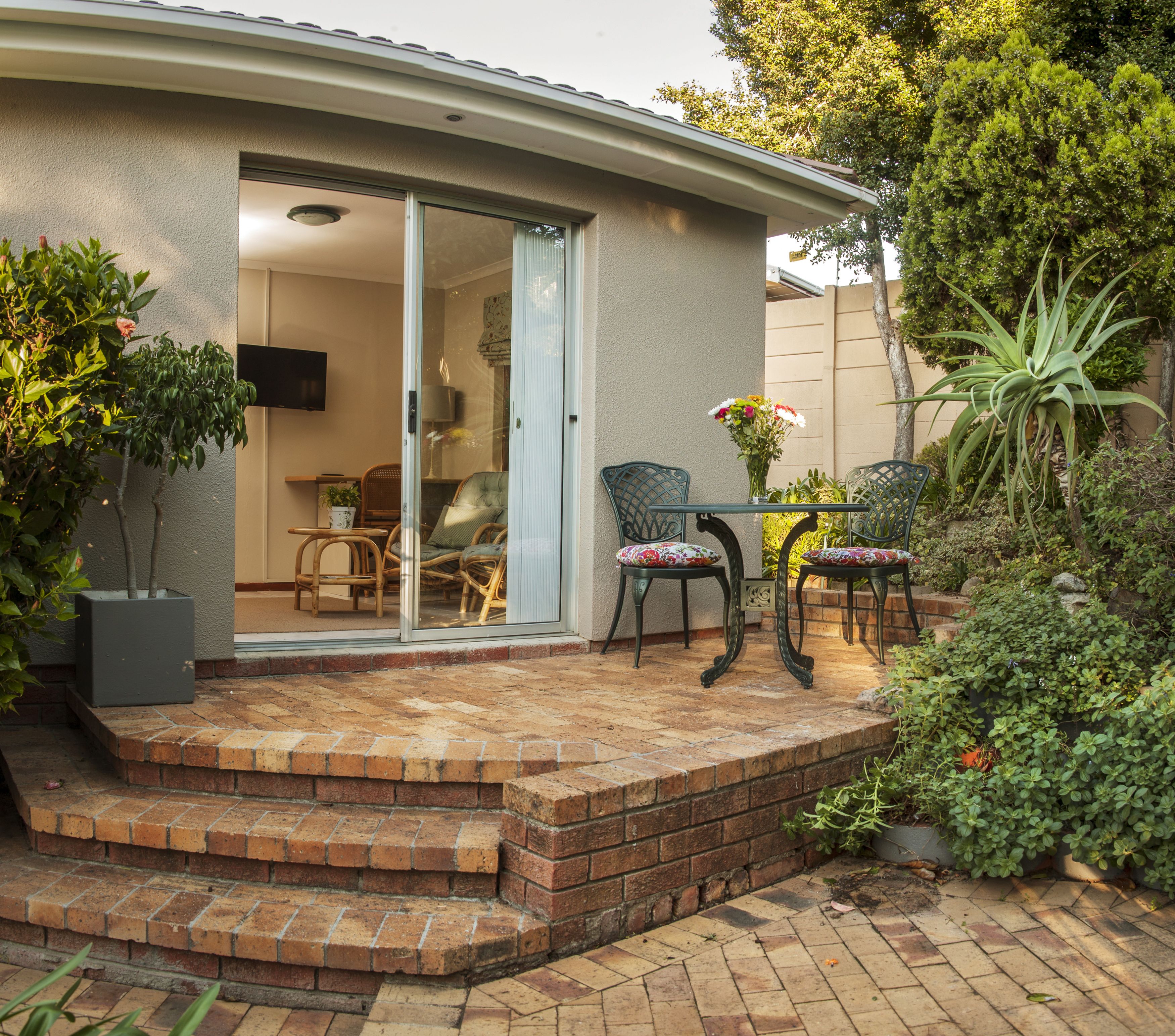 A patio with a brick floor, two wicker chairs, a small table, and a glass door leading to an interior space with a TV.