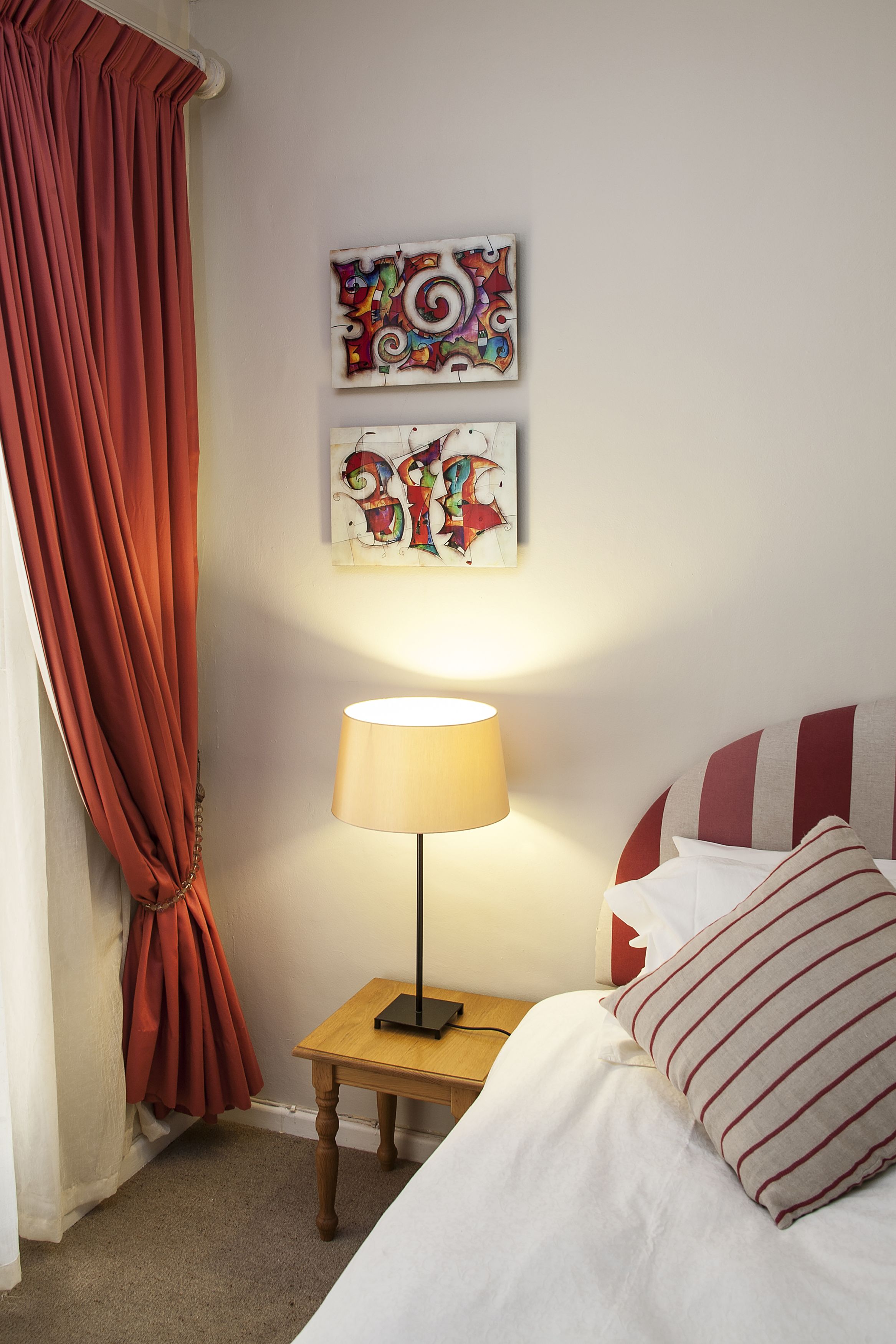 A neatly made bed with striped pillows, a small bedside table with a lamp, and two pieces of wall art.