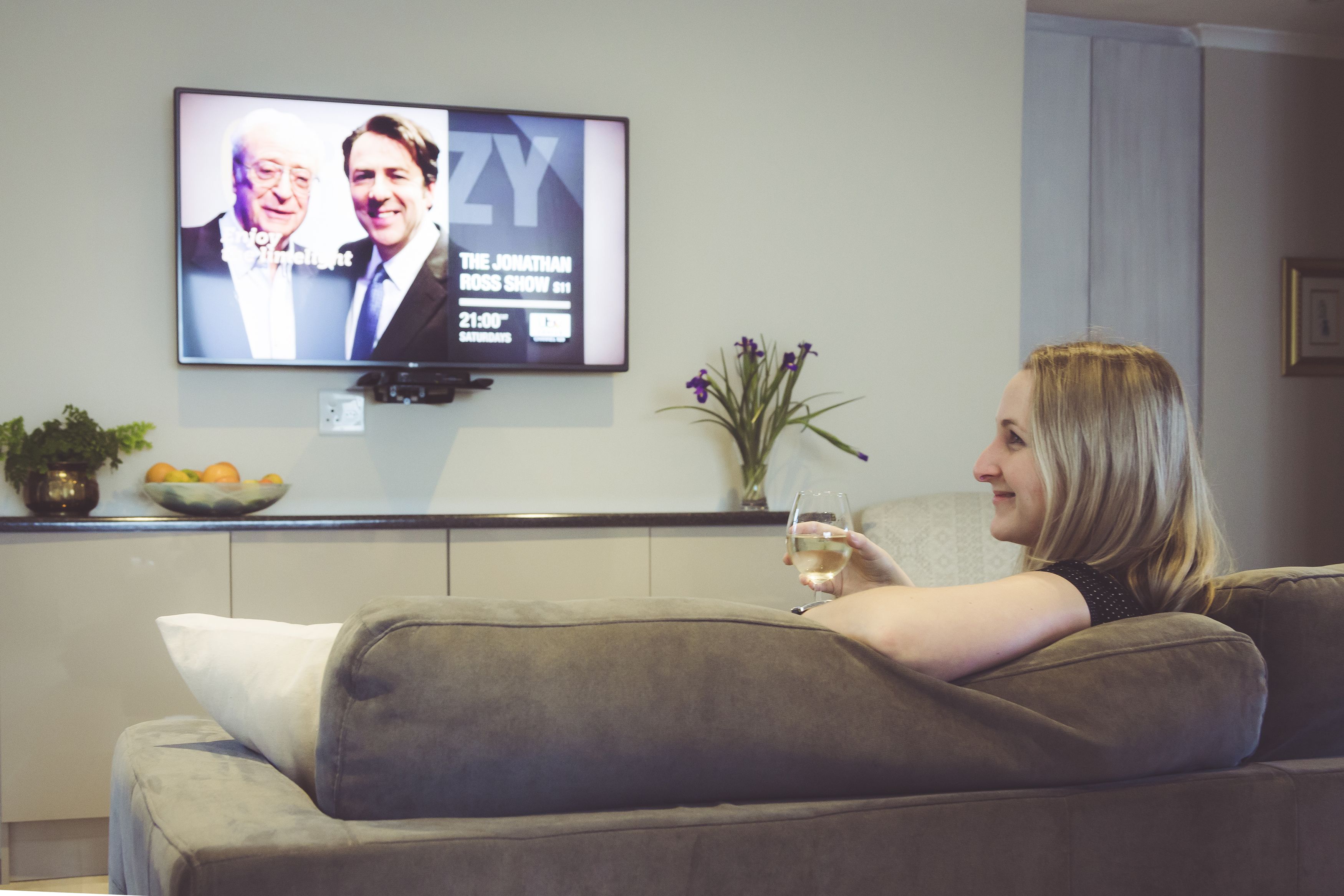 A person sitting on a grey couch, holding a glass of wine, with a TV displaying a show in the background.