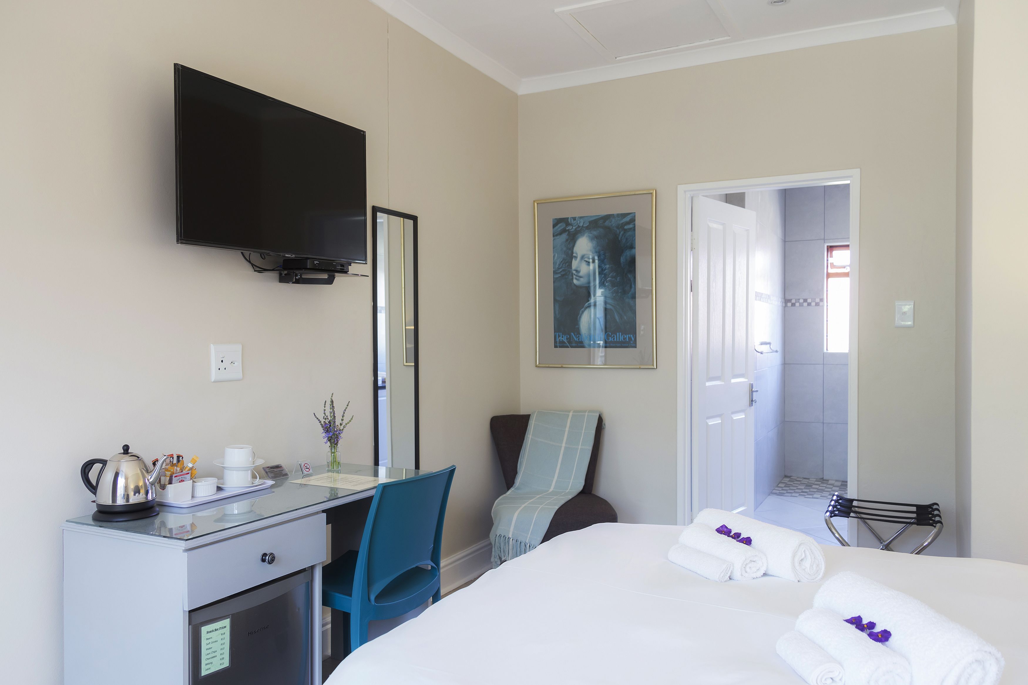A hotel room with a bed, a desk with a kettle, a chair, a television, and a shower area.
