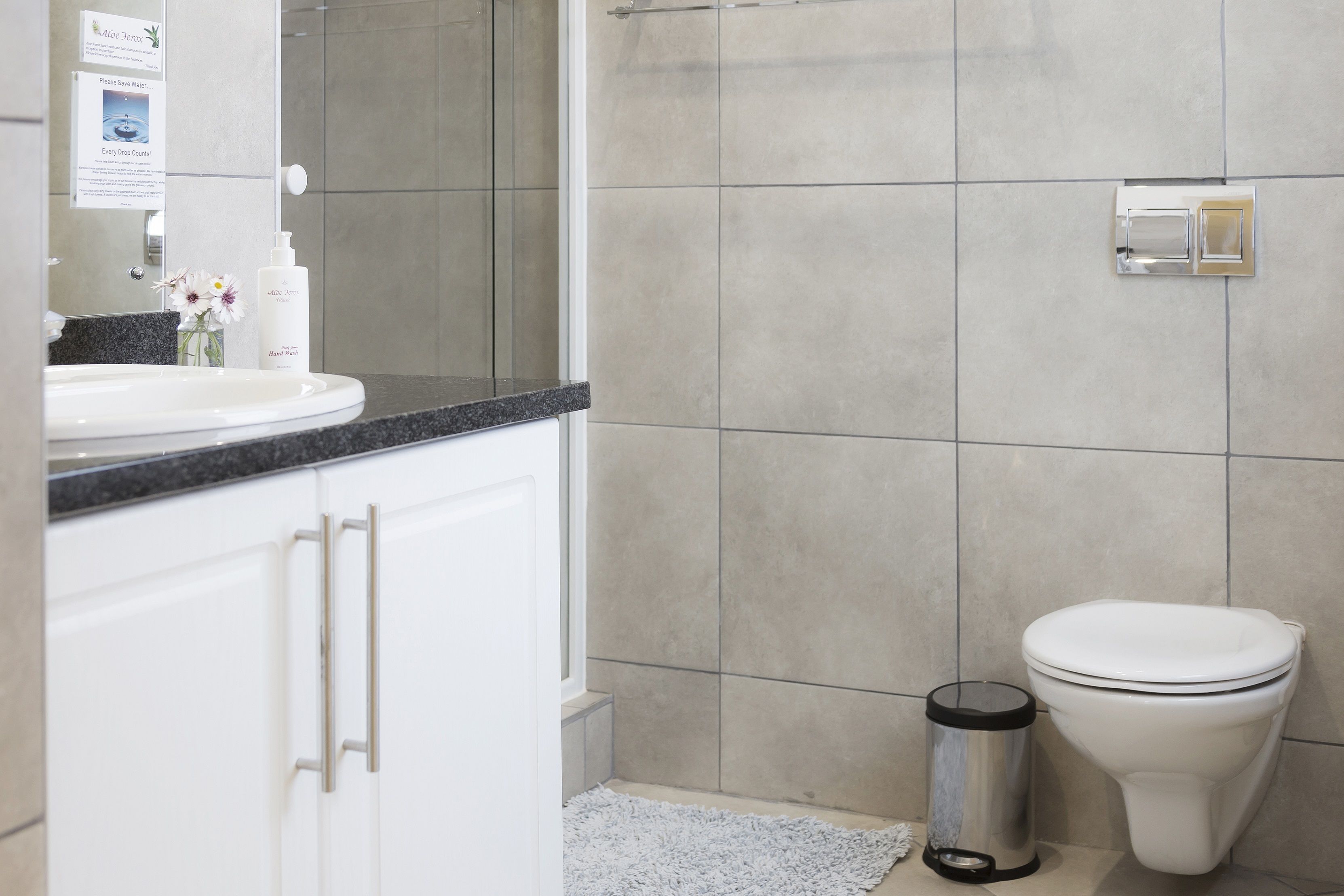 Une salle de bain avec un lavabo blanc, un miroir et des toilettes. On y trouve une poubelle et un petit tapis sur le sol.