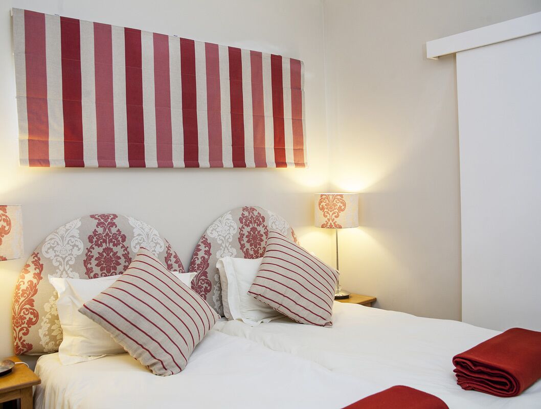 A neatly made bed with striped pillows, a red blanket, and a red towel on the side. There are two bedside lamps with patterned lampshades.