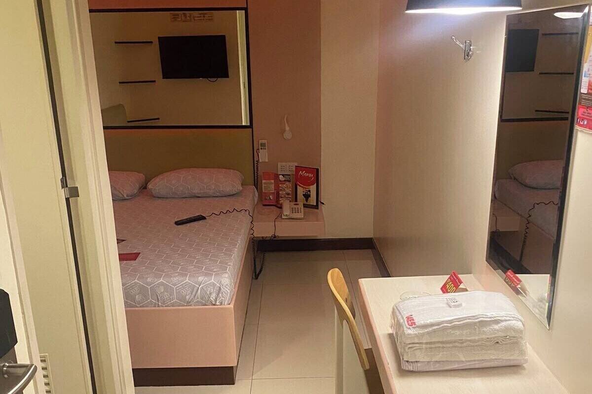 A compact hotel room with a bed, bedside table, mirror, and a small desk with a chair.