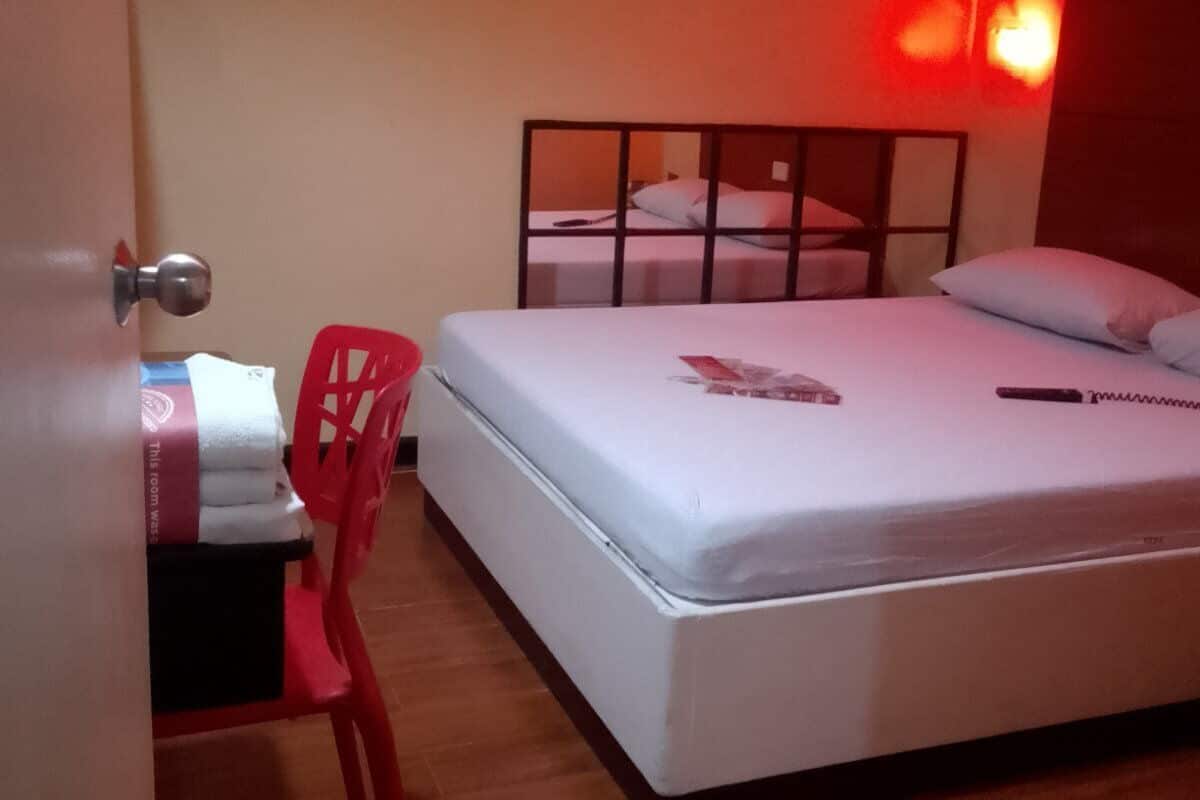 A single bed with a headboard, a red chair, a small table with a towel, and an air conditioning unit on the wall.
