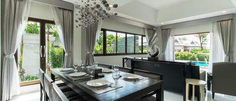 A modern dining area with a dark wooden table set for four, large windows with curtains, and a view of greenery outside.