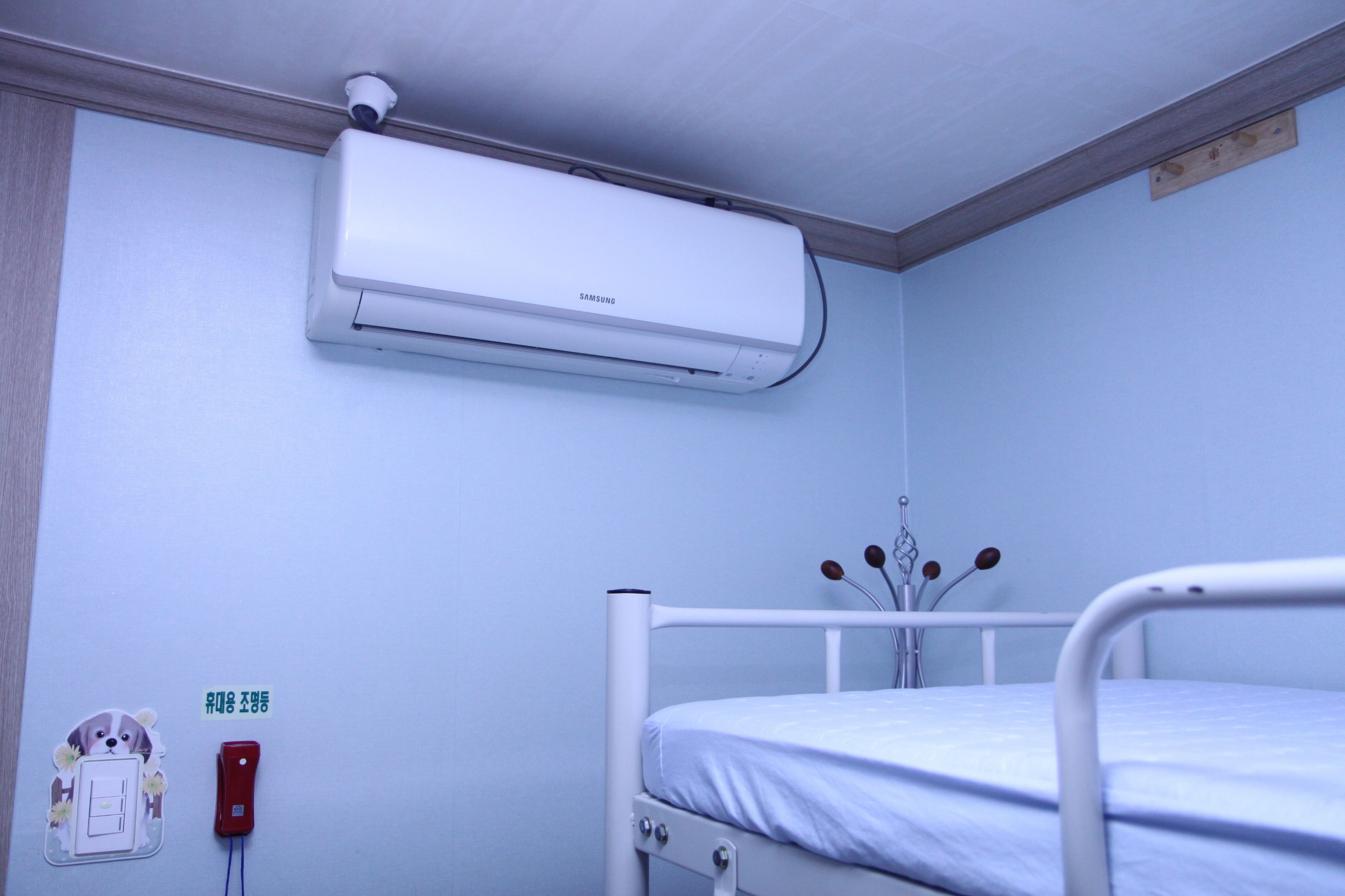 Quarto individual com uma máquina de ar condicionado branca, um telefone de emergência vermelho e uma câmera de segurança fixada na parede.