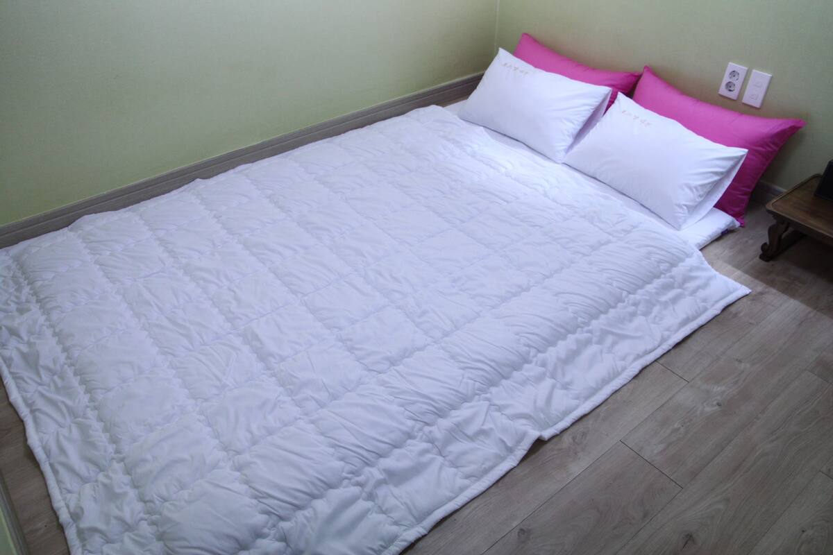 Ein Bett mit weißer Steppdecke und zwei Kissen, eines weiß und eines rosa, in einem Zimmer mit hellgrünen Wänden und Holzböden.