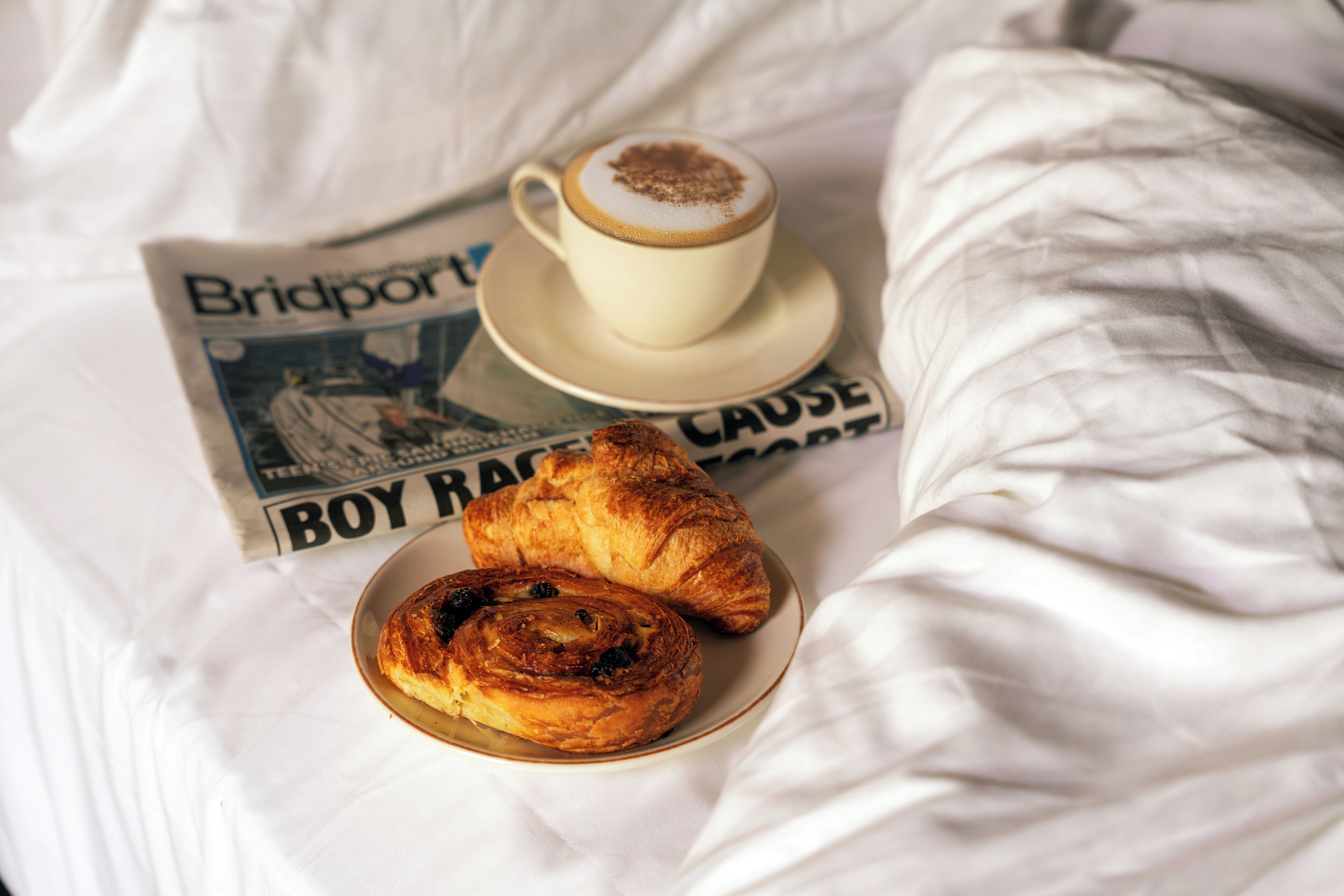 Una cama con un periódico, una taza de café y un croissant.