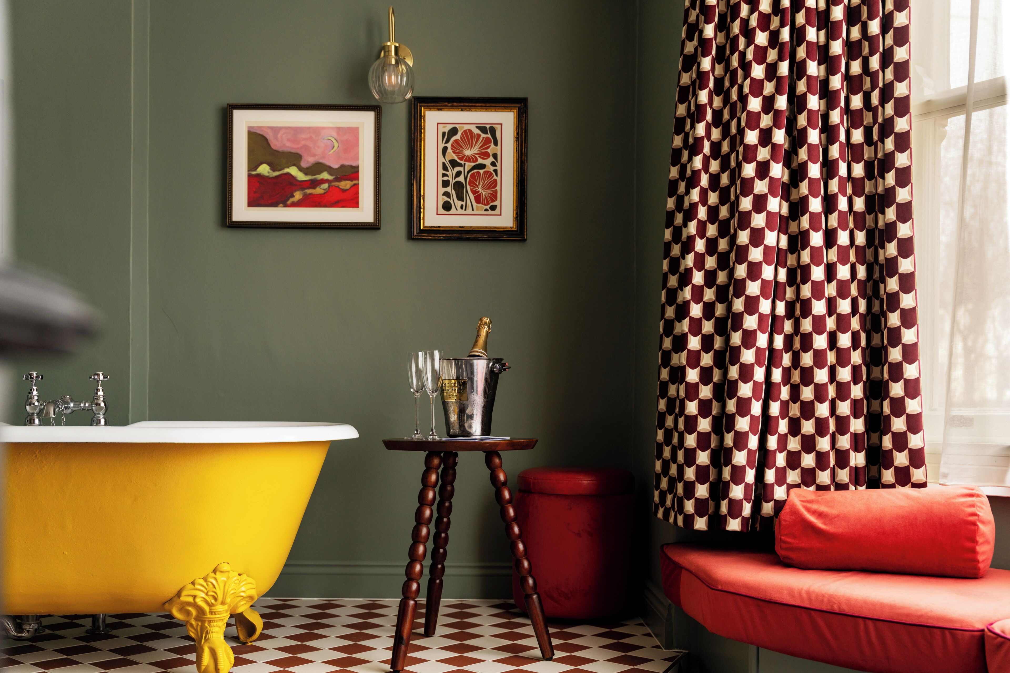 A bathroom with a yellow clawfoot bathtub, a red bench, and two framed artworks on the wall.