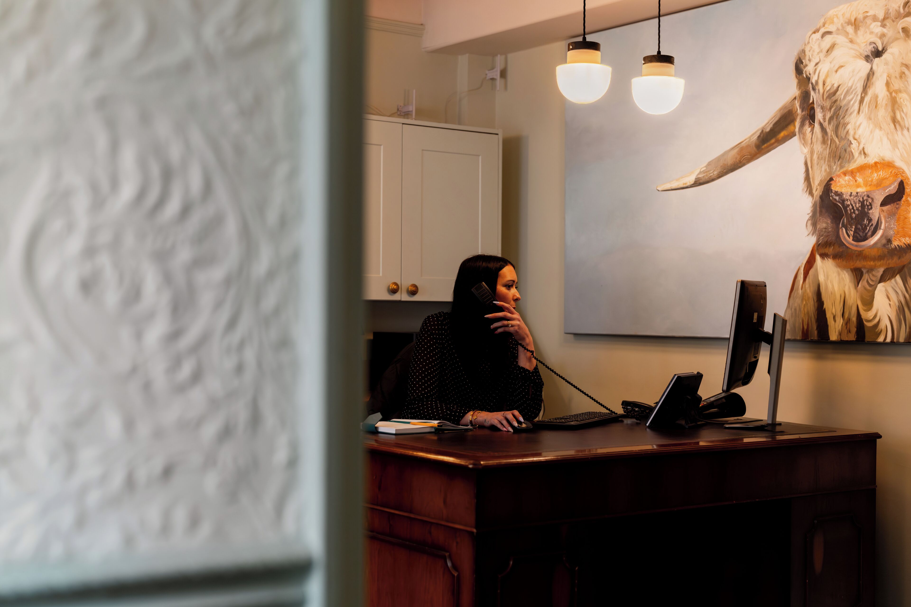 Une personne assise à un bureau, devant un ordinateur et un téléphone, avec un tableau représentant un taureau accroché au mur.