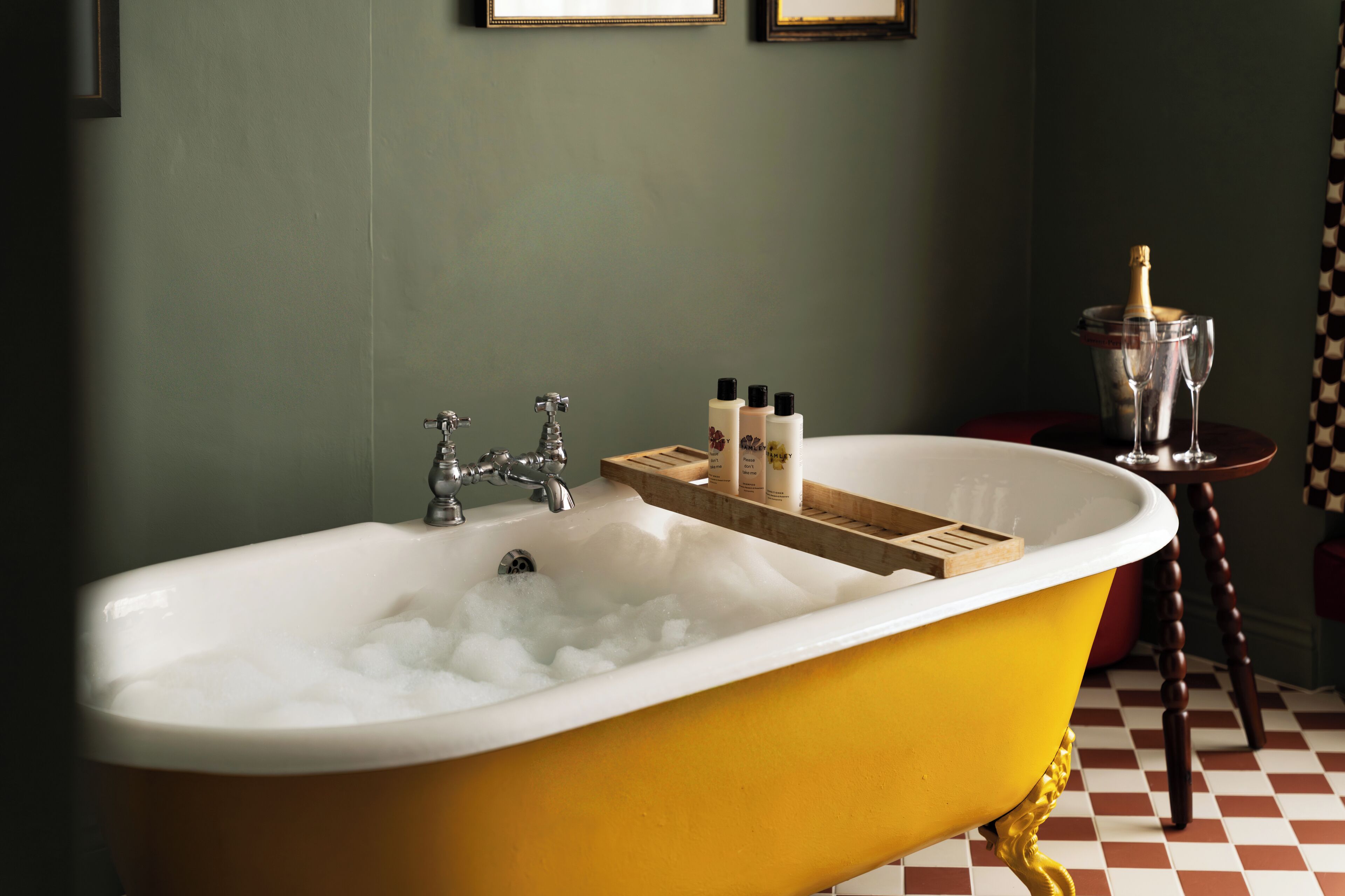 A yellow bathtub with a wooden tray holding toiletries, a bottle of champagne, and two glasses on a checkered floor.
