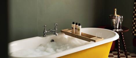 A yellow bathtub with a wooden tray holding toiletries, a bottle of champagne, and two glasses on a checkered floor.