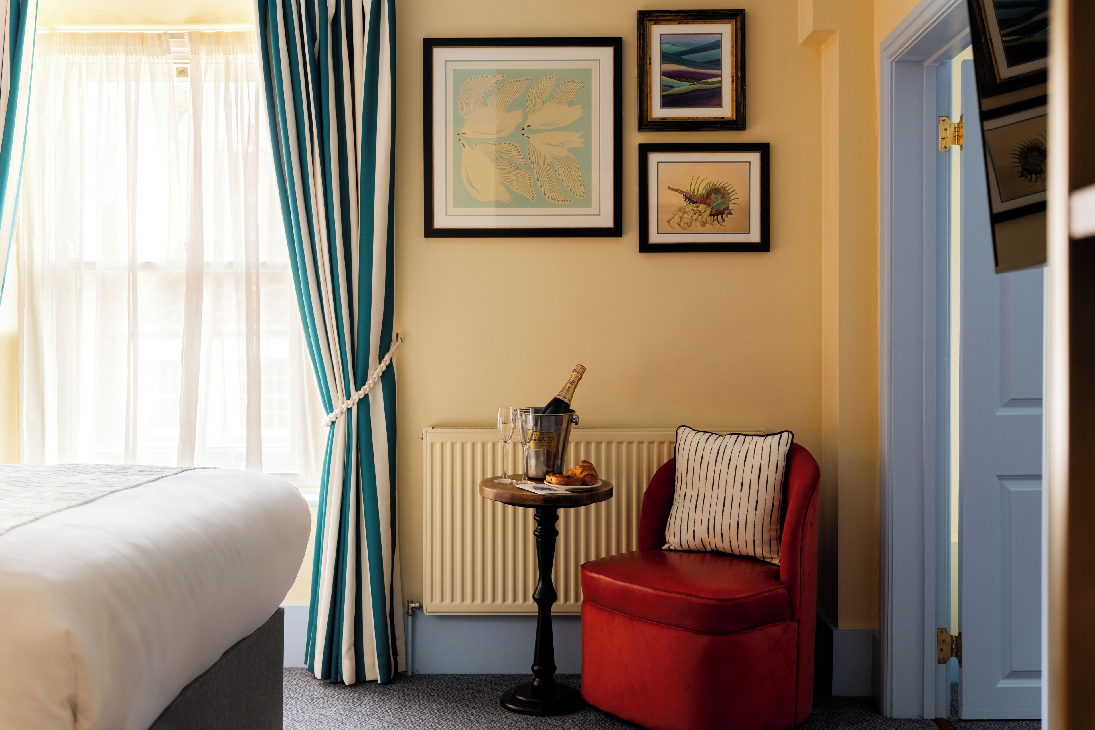 Habitación de hotel con una cama, un sillón rojo, una mesita con una botella y vasos, y cuadros en la pared.