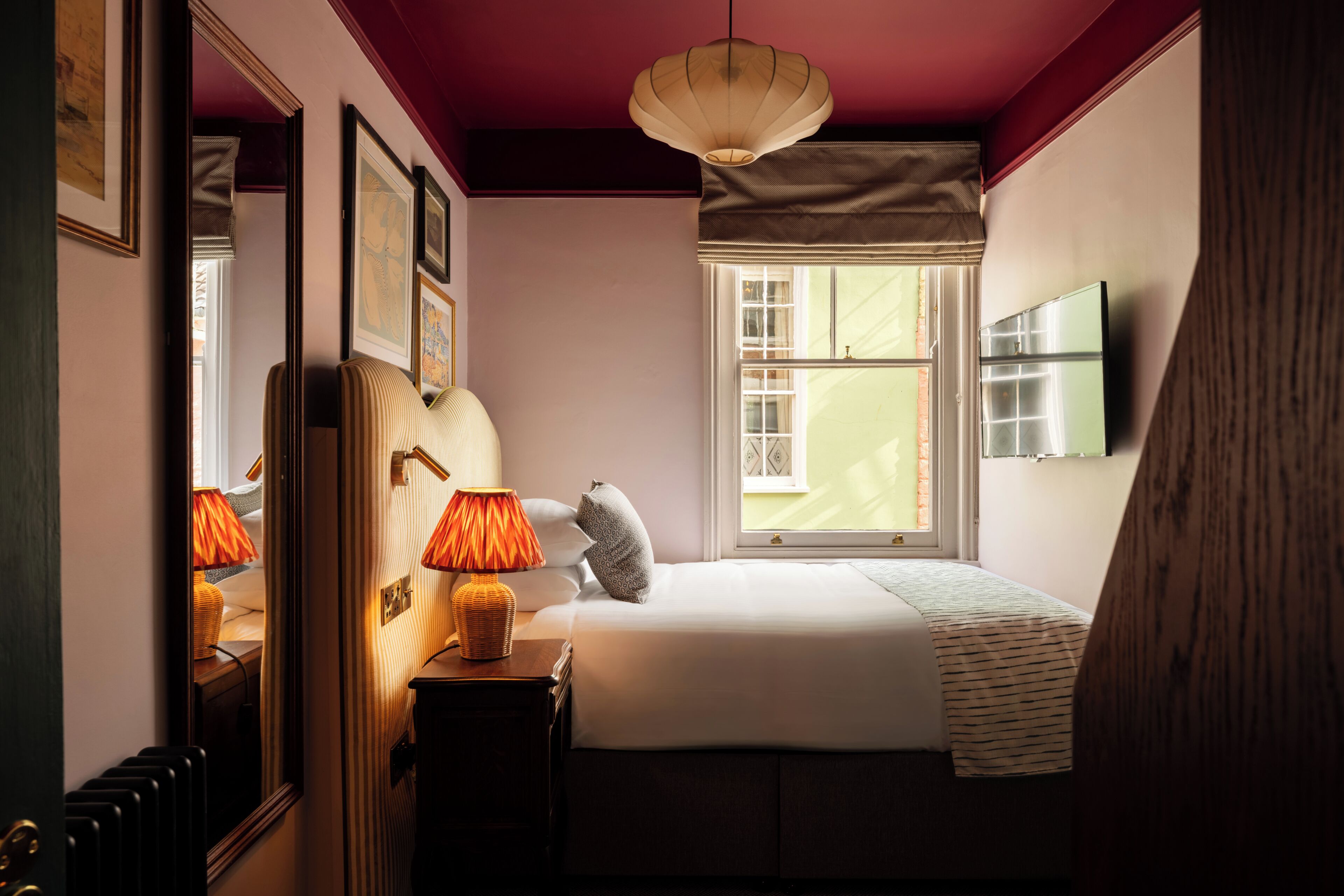 A bedroom with a bed, a bedside lamp, a window, and a mirror.
