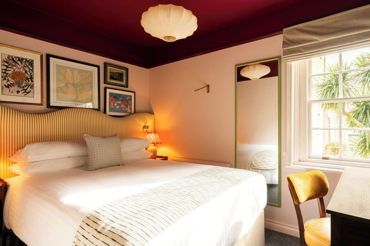 A bedroom with a bed, bedside table, lamp, chair, window, and artwork on the wall.