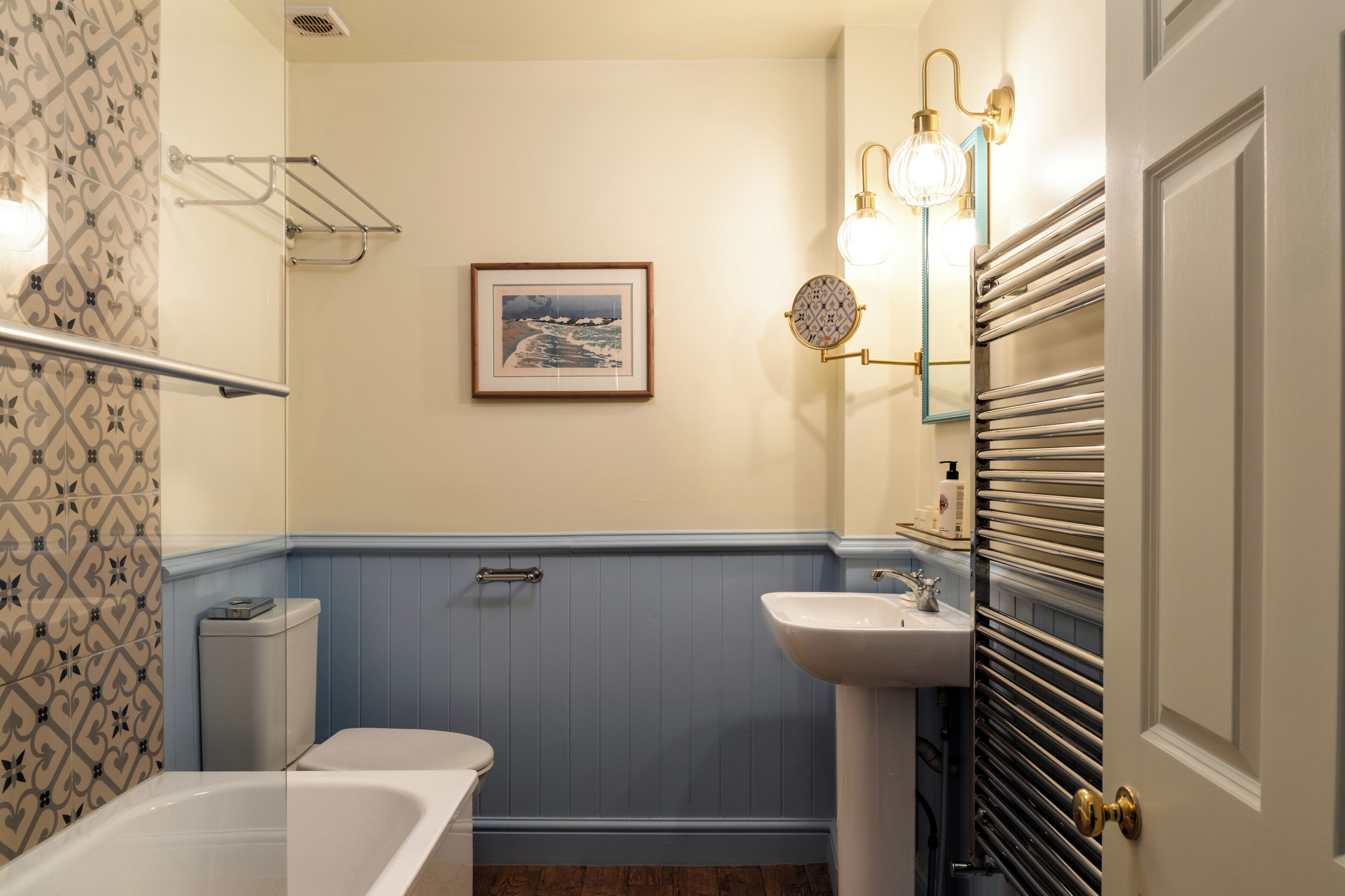 Een badkamer met een bad, wastafel, toilet, handdoekhouder, een ingelijst schilderij aan de muur en een spiegel met verlichting.