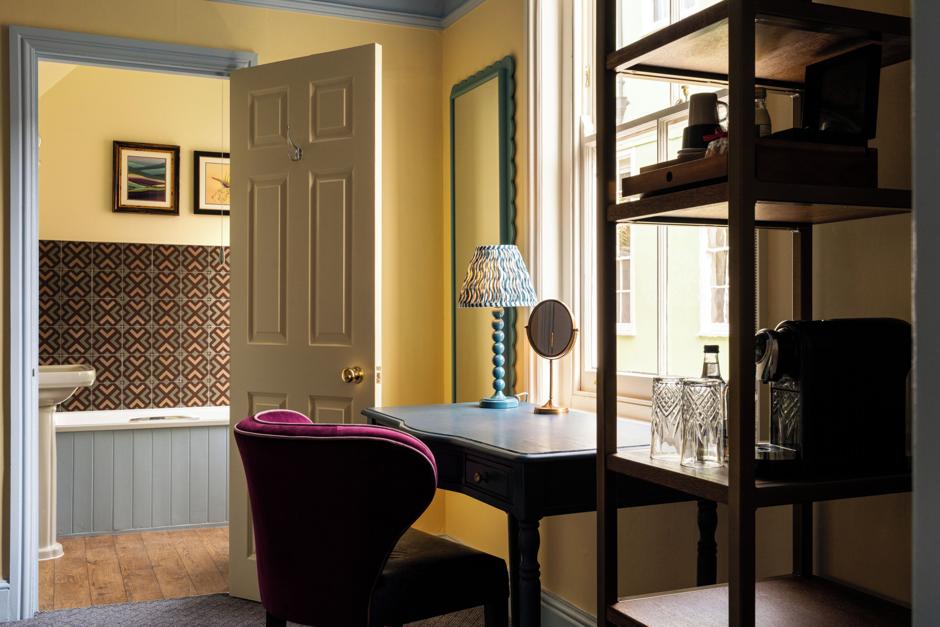 A room with a desk, chair, mirror, and shelves with glassware.