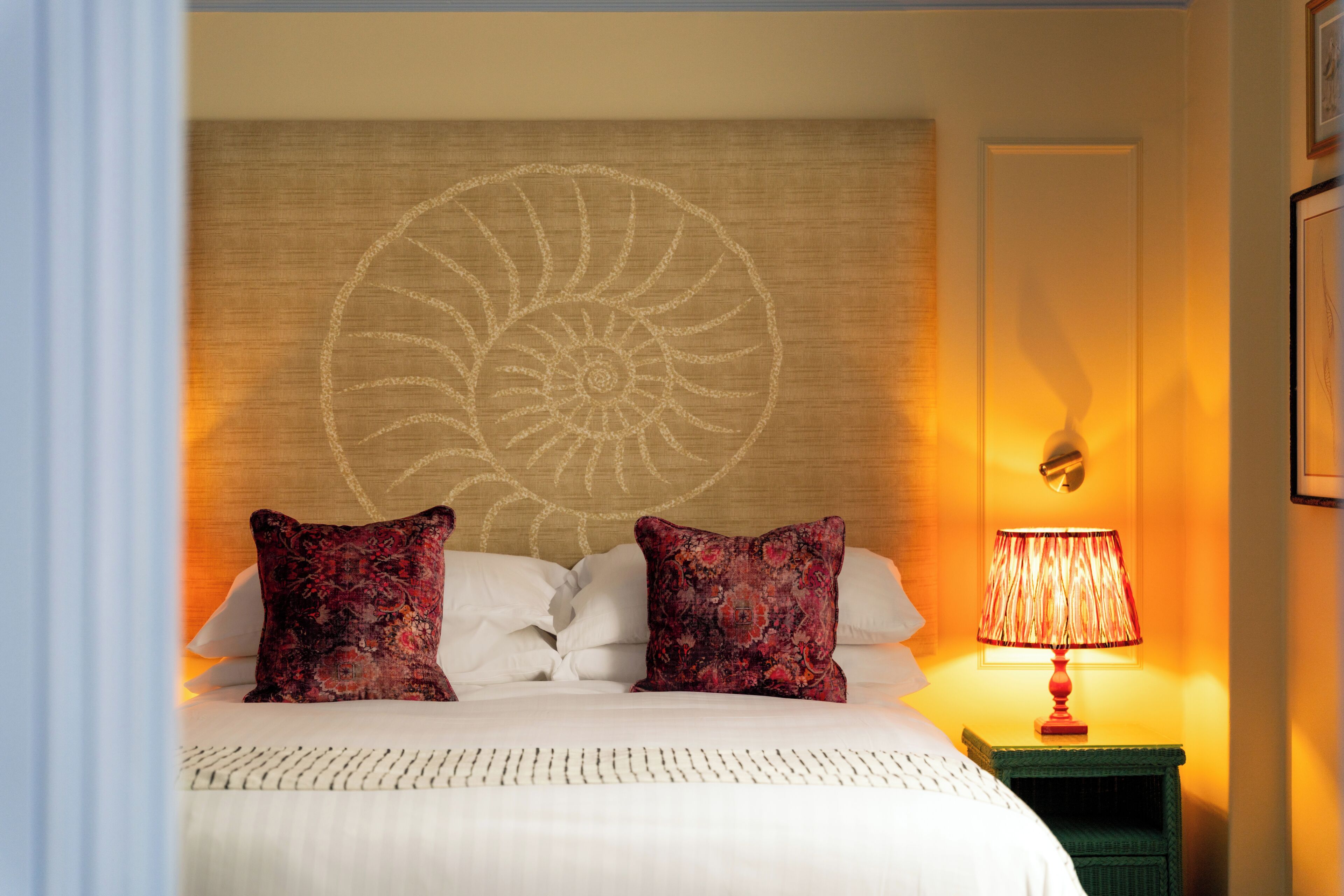 Una habitación de hotel con una cama, dos almohadones decorativos, un tapiz en la pared, una mesita de noche con una lámpara y un cuadro enmarcado.