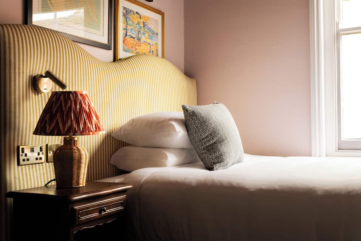 A neatly made bed with pillows, a bedside lamp, a wall socket, and a framed picture above the bed.