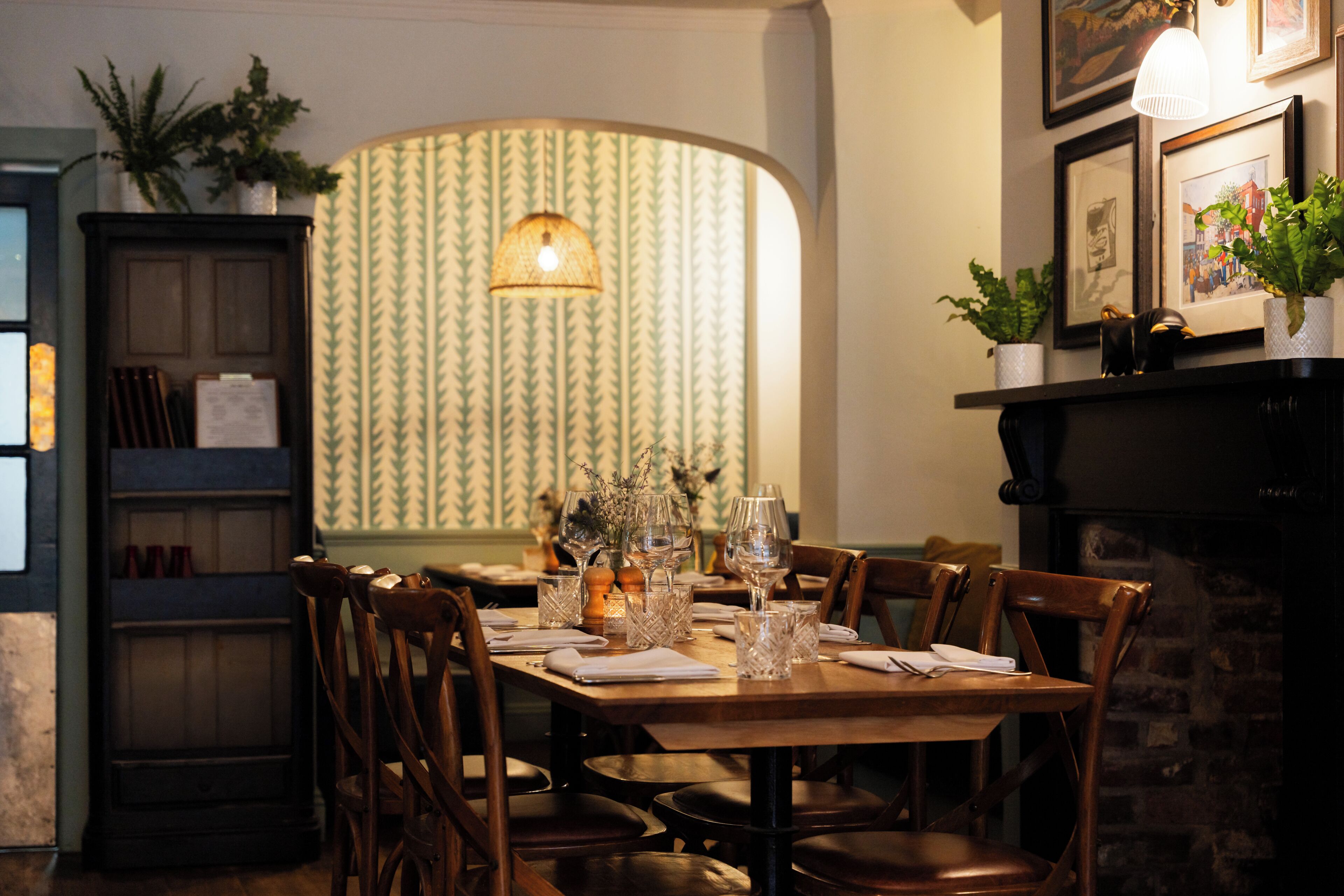 A dining area with a wooden table set for four, surrounded by chairs, with a patterned wallpaper and a hanging light fixture above.