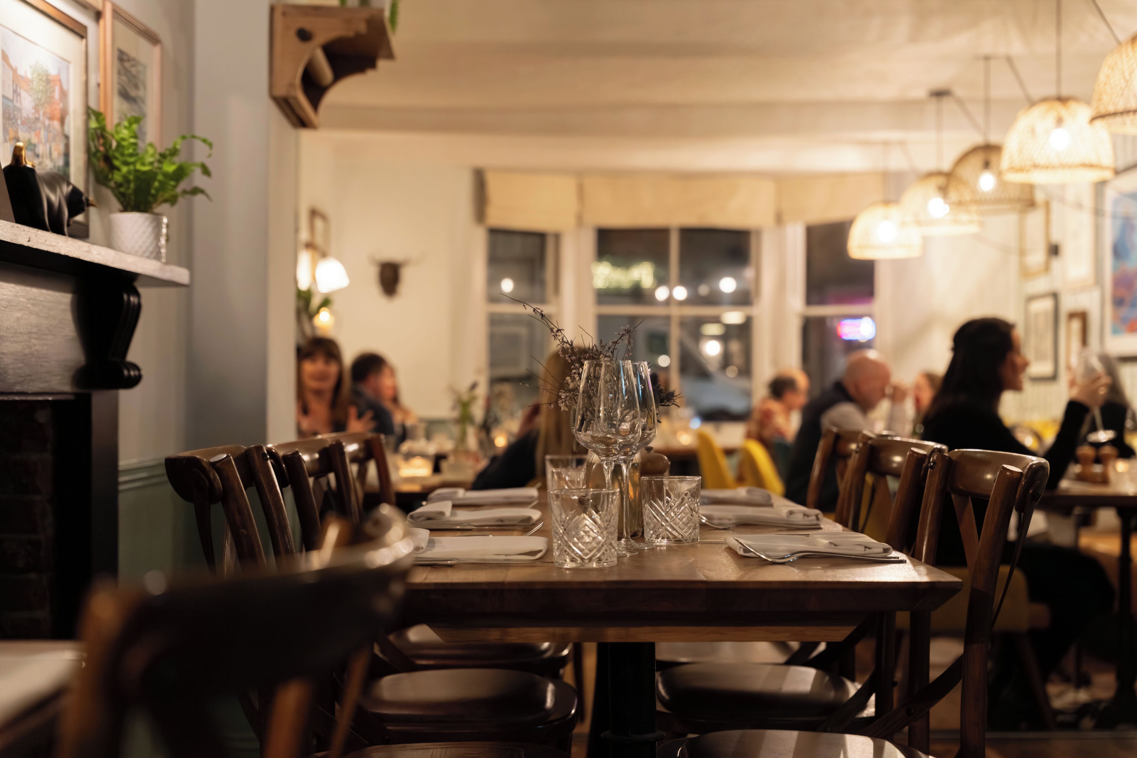 Interior de restaurante con mesas y sillas de madera, un jarrón con ramas y cuadros en la pared.