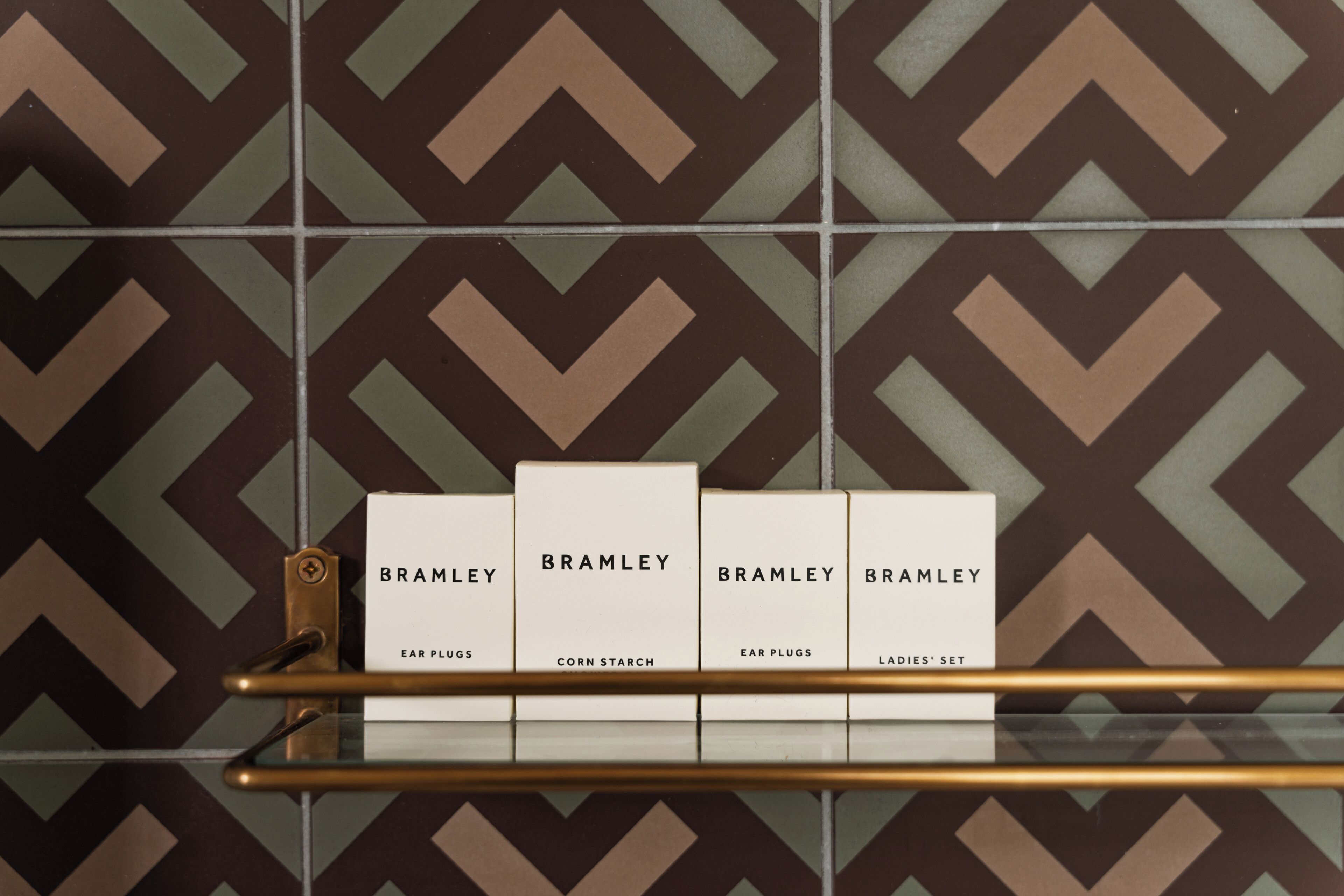 Una pared con baldosas y un diseño geométrico, con un estante metálico que sostiene cuatro cajas etiquetadas 'BRAMLEY' con diferentes nombres de productos.