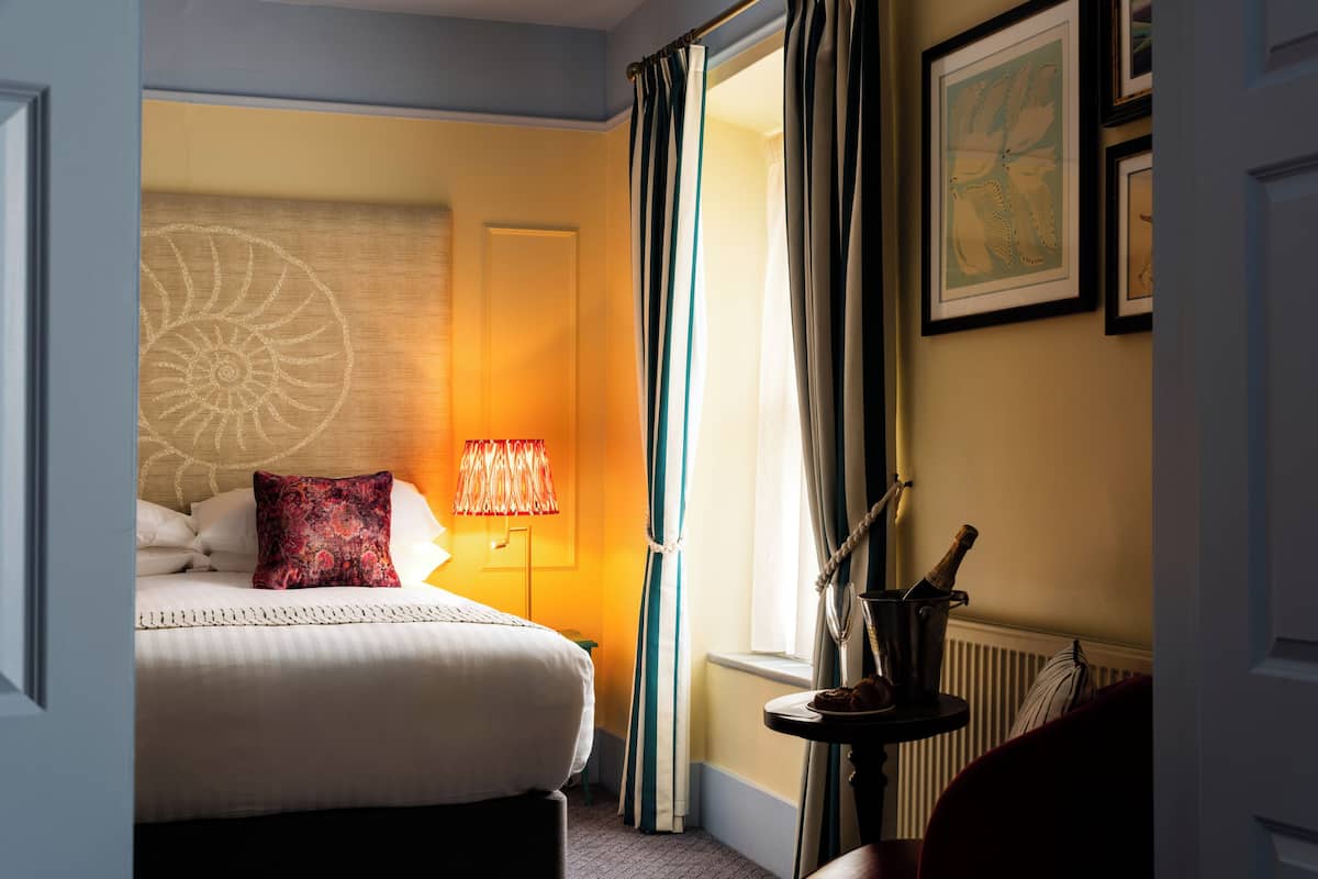 A hotel room with a bed, a lamp, curtains, a framed picture, and a bottle on a side table.