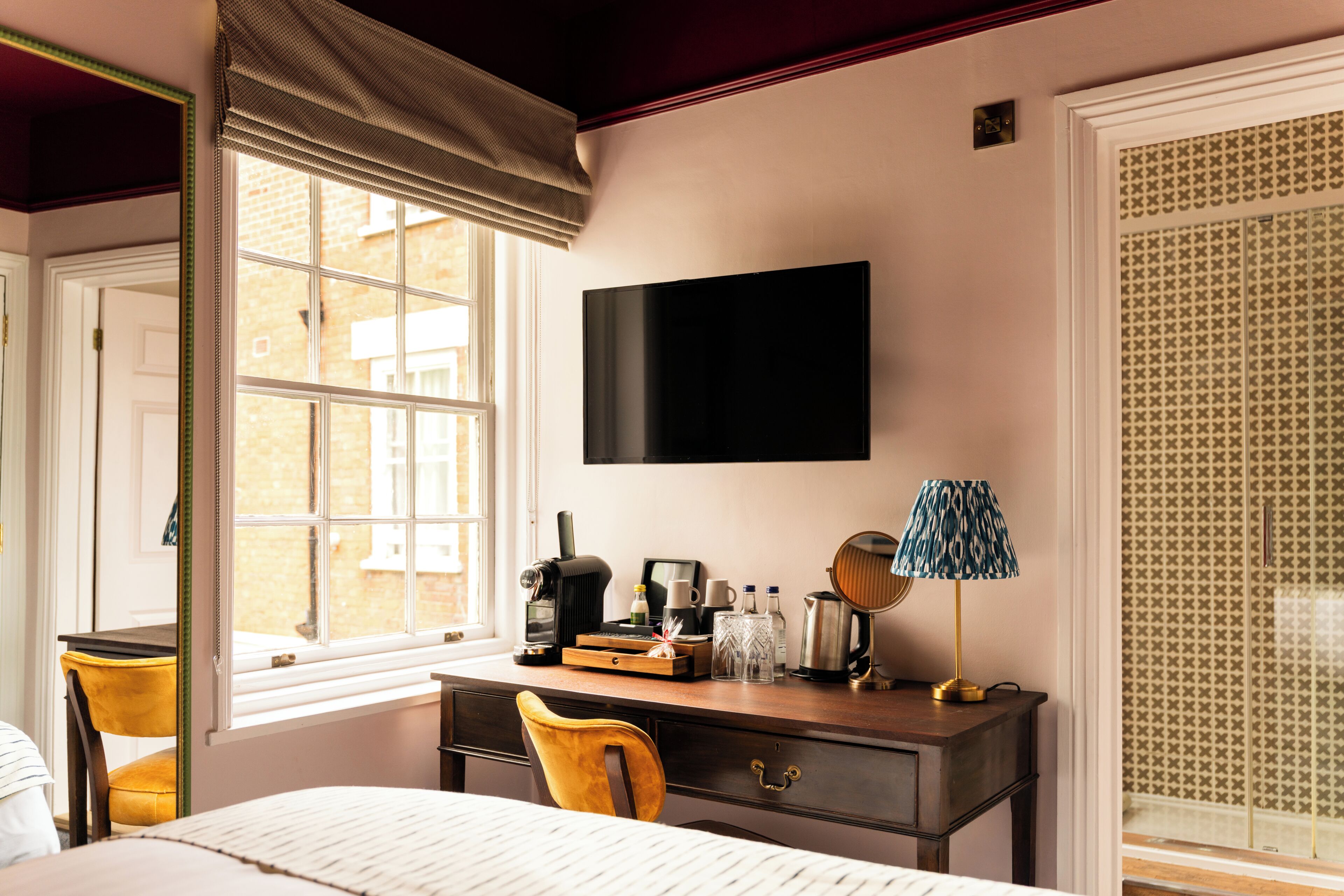 A hotel room with a bed, a desk with a flat-screen TV, a window with a view of buildings, a chair, and a patterned wall.