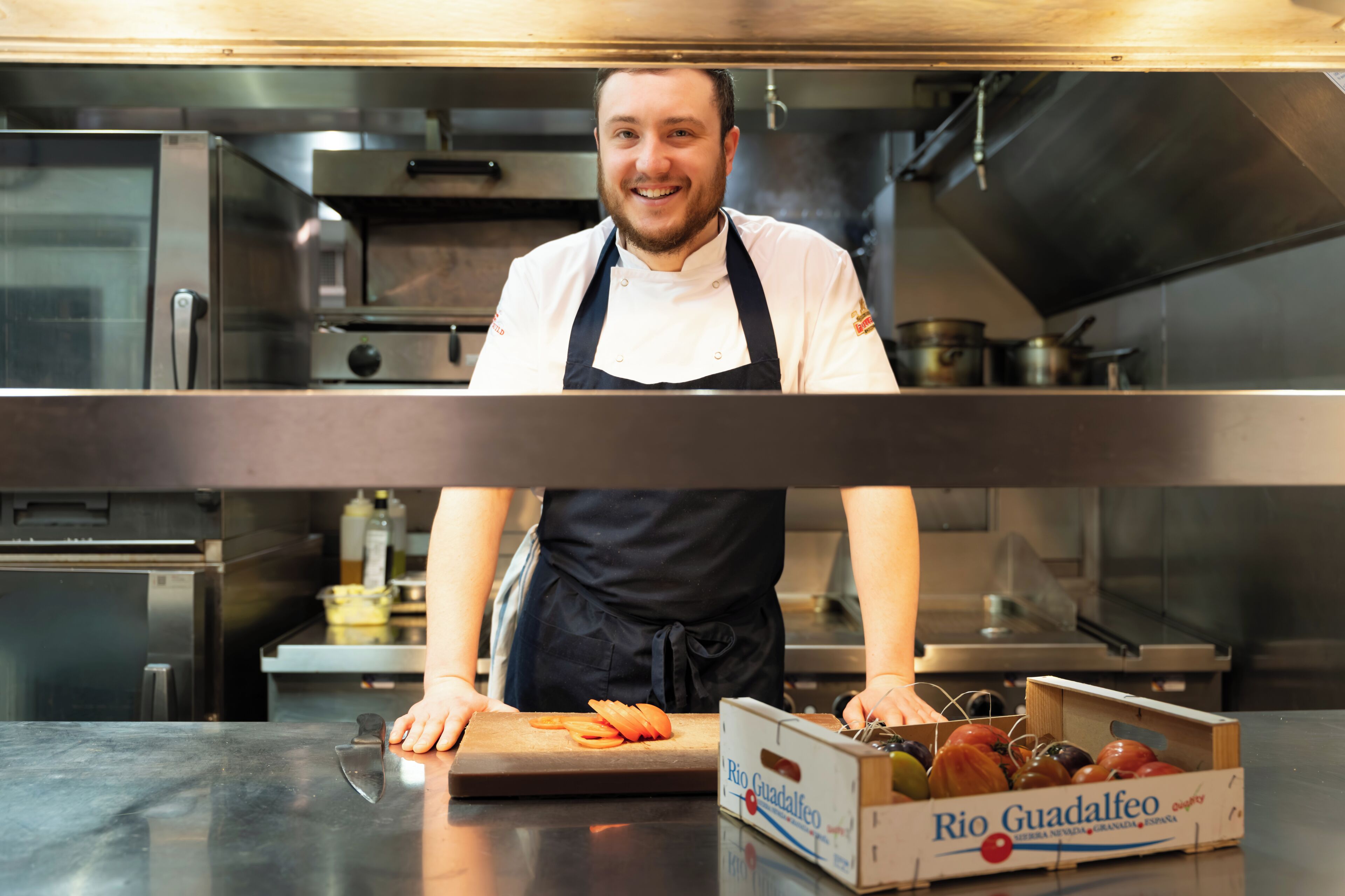 A chef in a professional kitchen with stainless steel appliances, a cutting board with vegetables, and a box of Rio Guadalfeo wine.