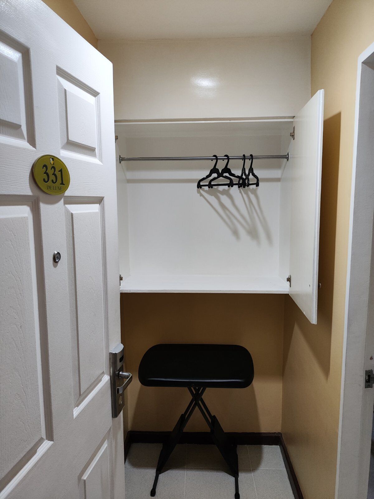 Een compacte kamer met een witte deur met het nummer 331, een klein zwart strijkplank en een witte garderobe met een metalen kledingroede.