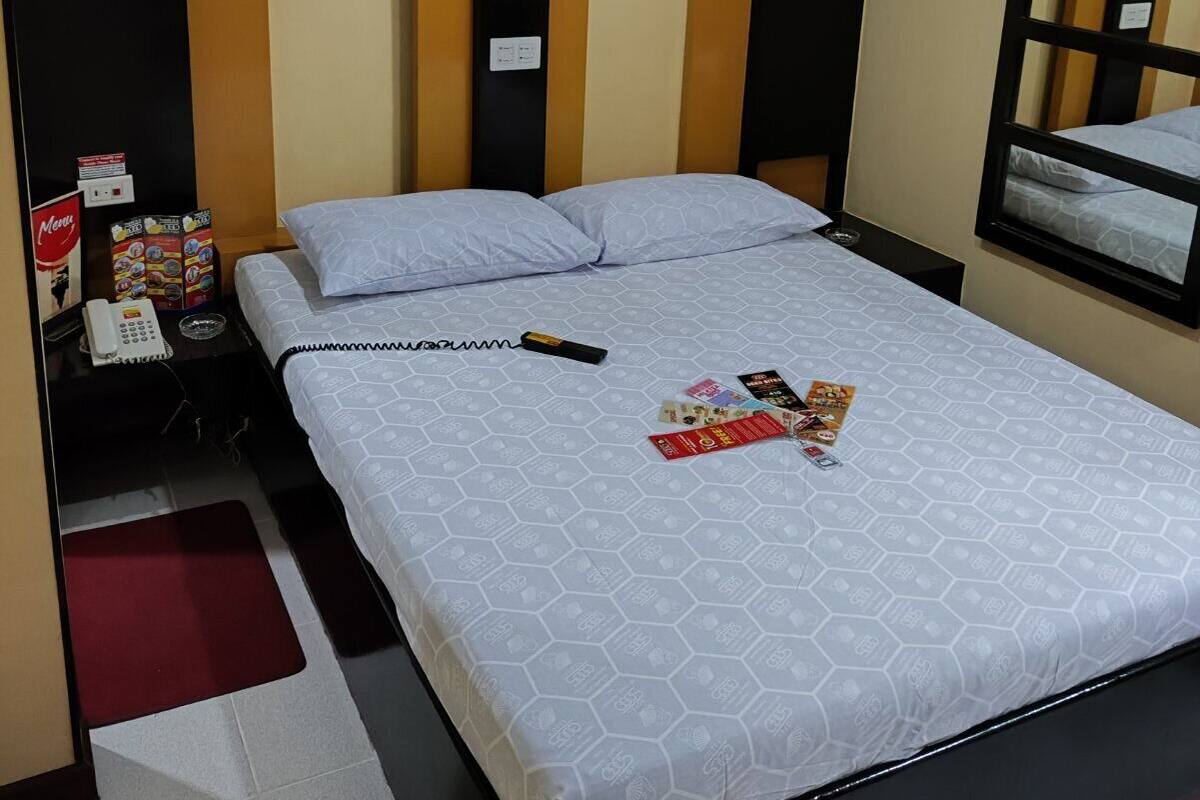 A hotel room with a bed, a bedside table with a phone and magazines, an air conditioner, and a window with a view.