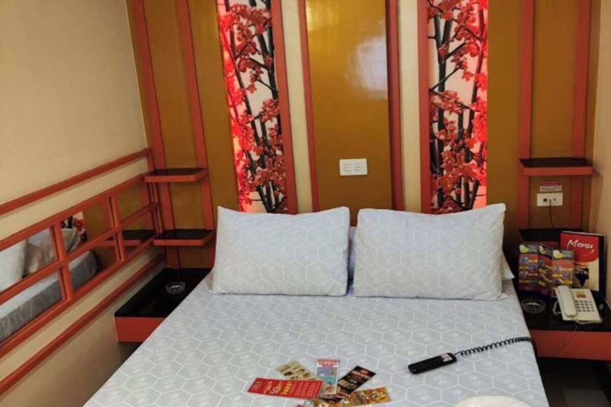 A hotel room with a bed, two pillows, a nightstand with a phone, and a decorative panel with red flowers.