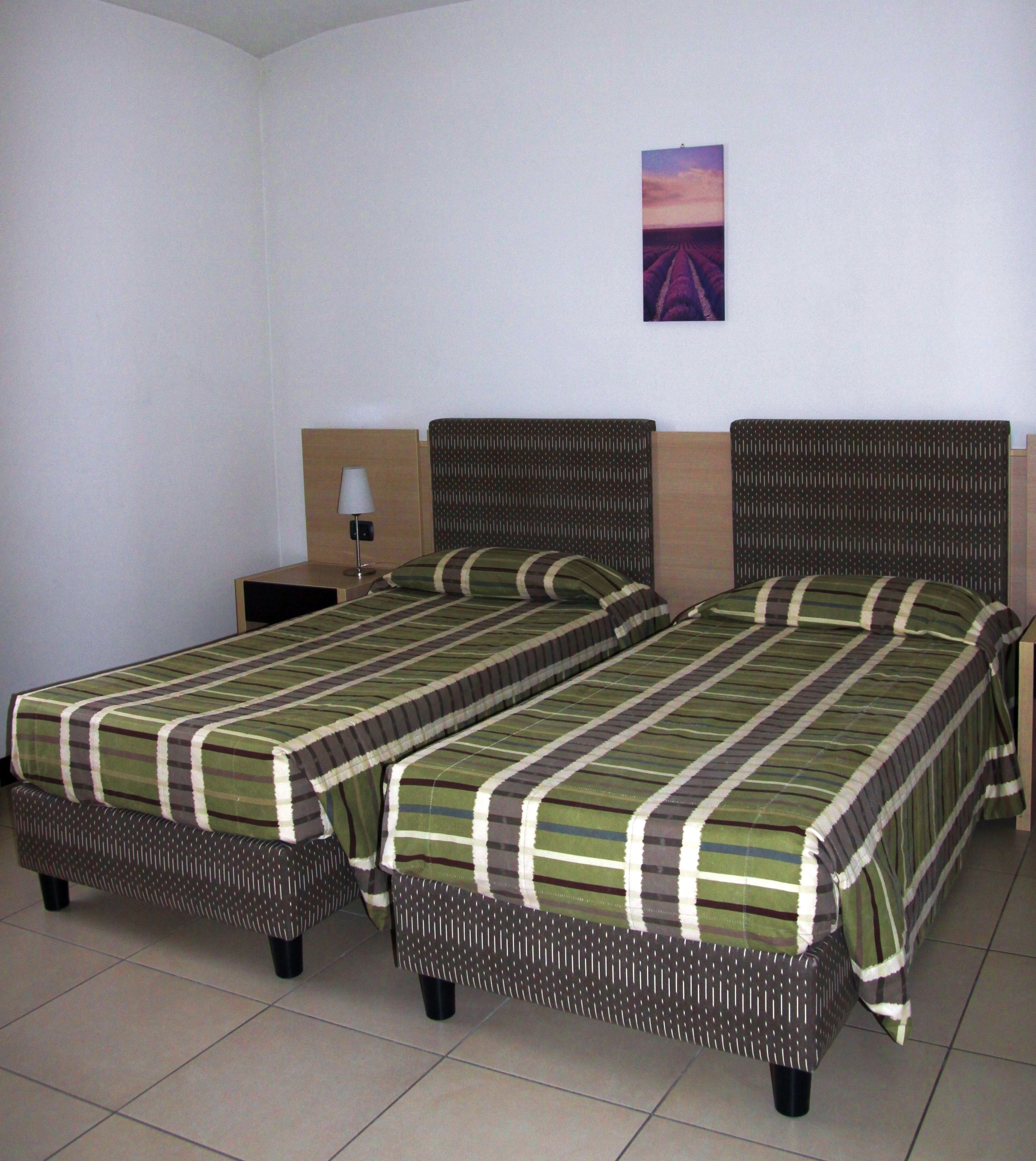 A hotel room with two single beds, a nightstand, a lamp, and a wall-mounted picture.