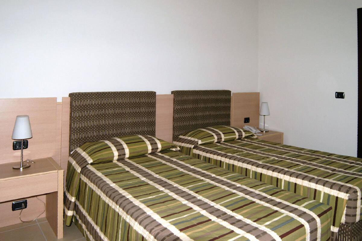 Habitación de hotel con dos camas individuales, cabecera de madera, mesita de noche con lámpara y toma de corriente.