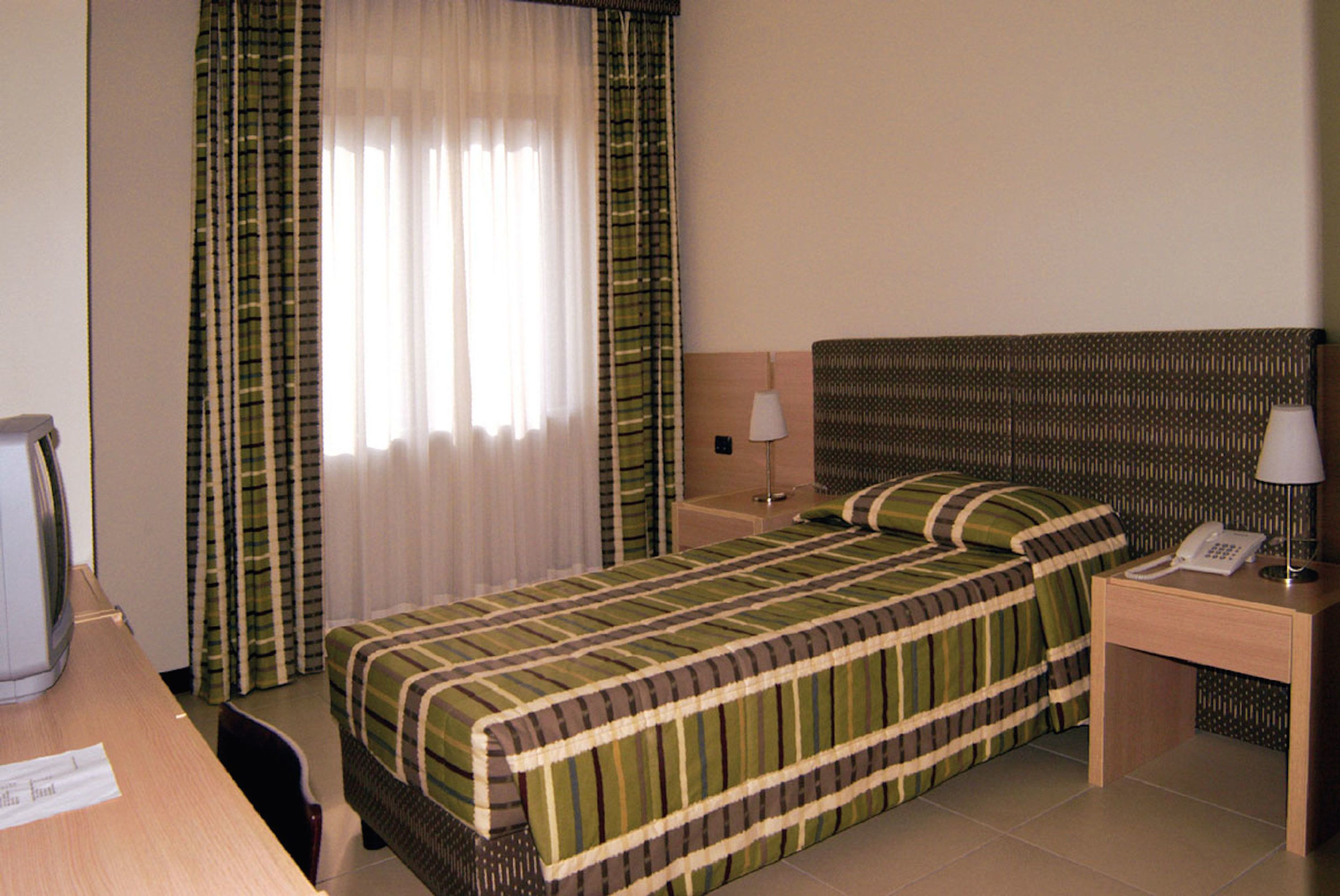 Una camera d'albergo con un letto, una scrivania con il telefono, una lampada e una finestra con tende.