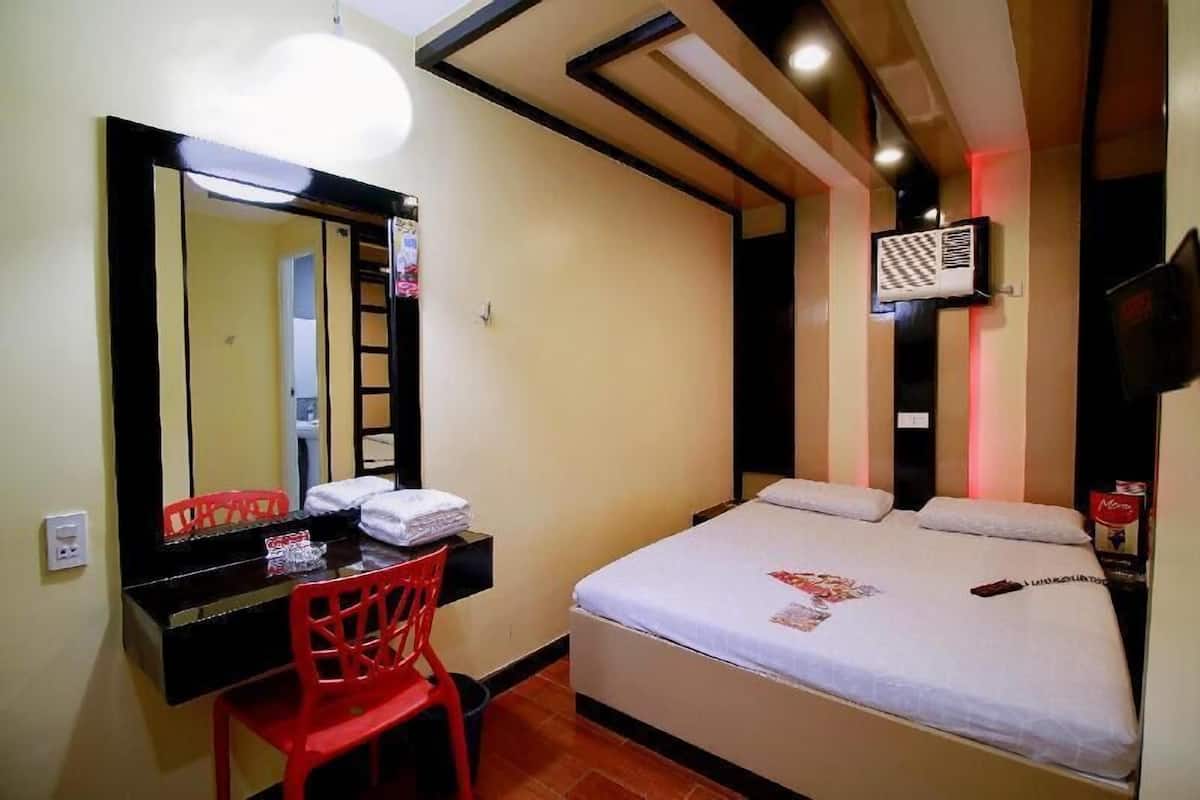 A hotel room with a bed, a desk with a chair, a mirror, a wall-mounted TV, and a ceiling light fixture.