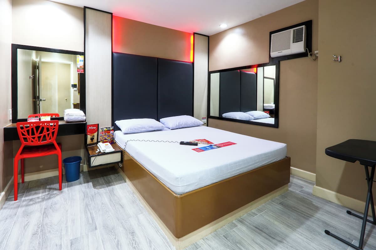 A hotel room with a large bed, a red chair, a small table, a trash bin, and a mirror.