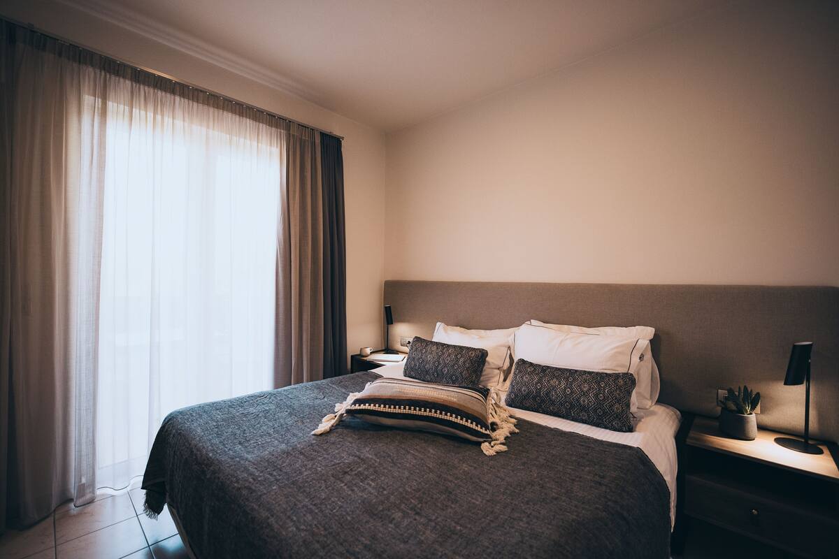 Uma cama bem arrumada com um cobertor escuro e almofadas estampadas, uma mesa de cabeceira com uma luminária e uma pequena planta, e uma janela grande com cortinas leves.