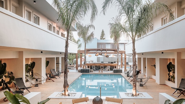 A pool area with lounge chairs, a wooden table, and a bar in the background. - Sentido More Meni Residence (Kos)