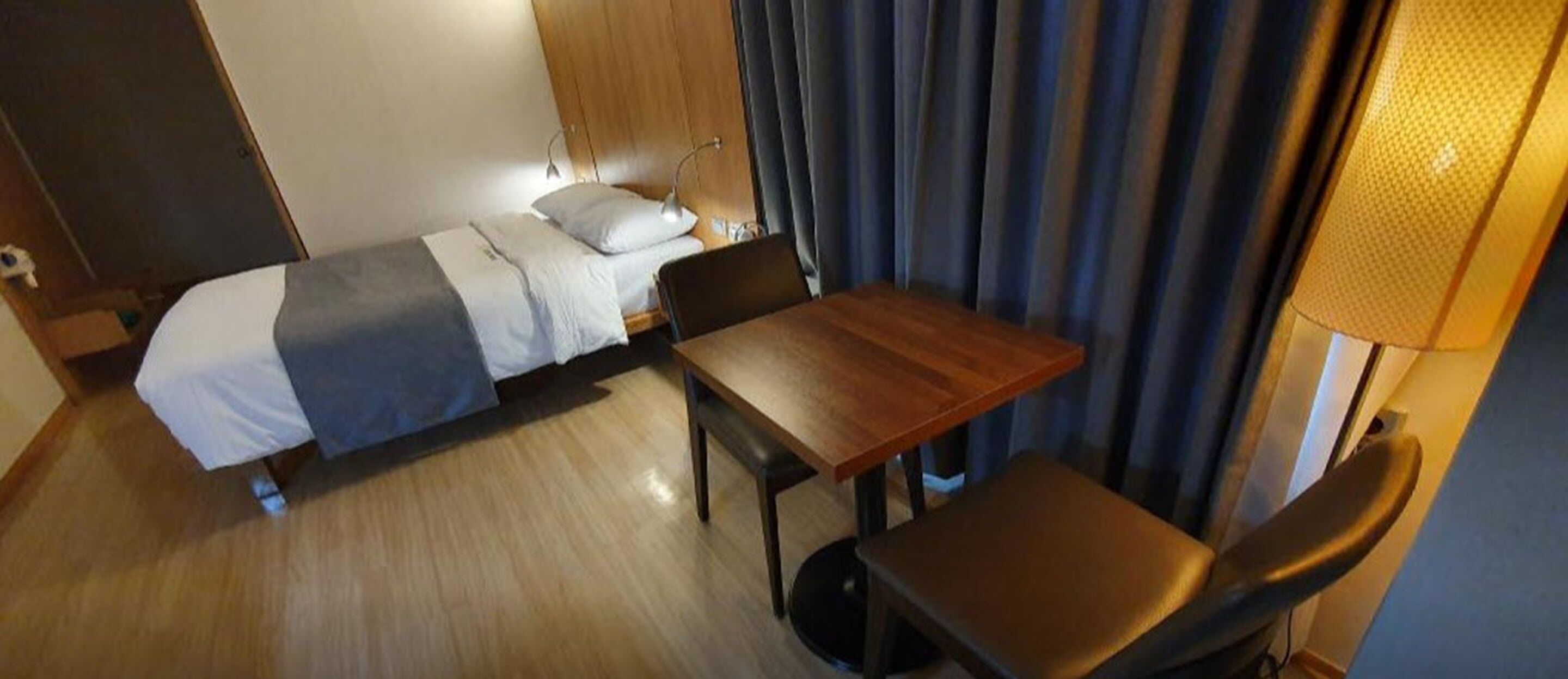 A hotel room with a bed, a wooden table, a chair, and a lamp.