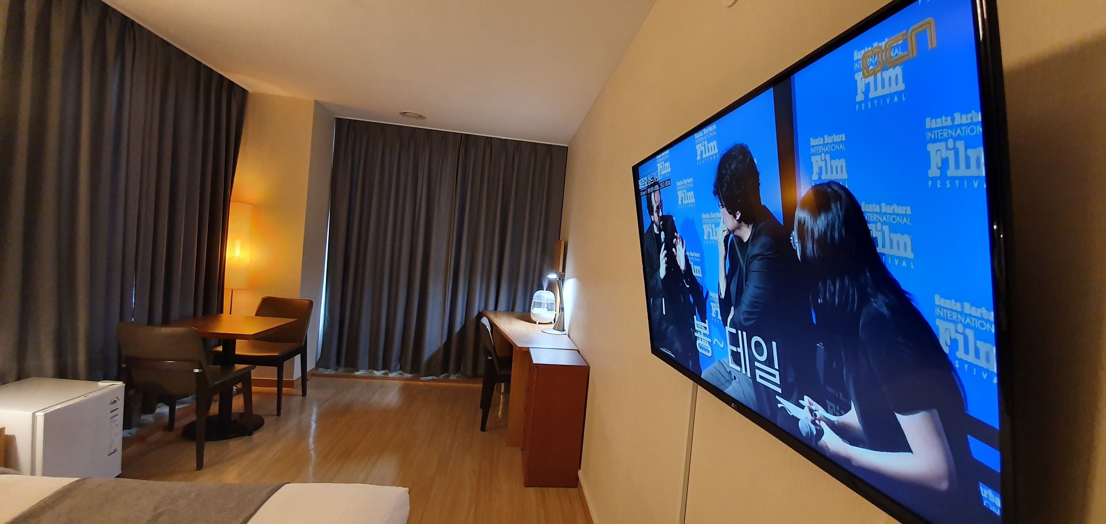 Habitación de hotel con un televisor de pantalla plana grande mostrando un festival de cine, una mesa de comedor de madera, un refrigerador blanco pequeño y un sofá.