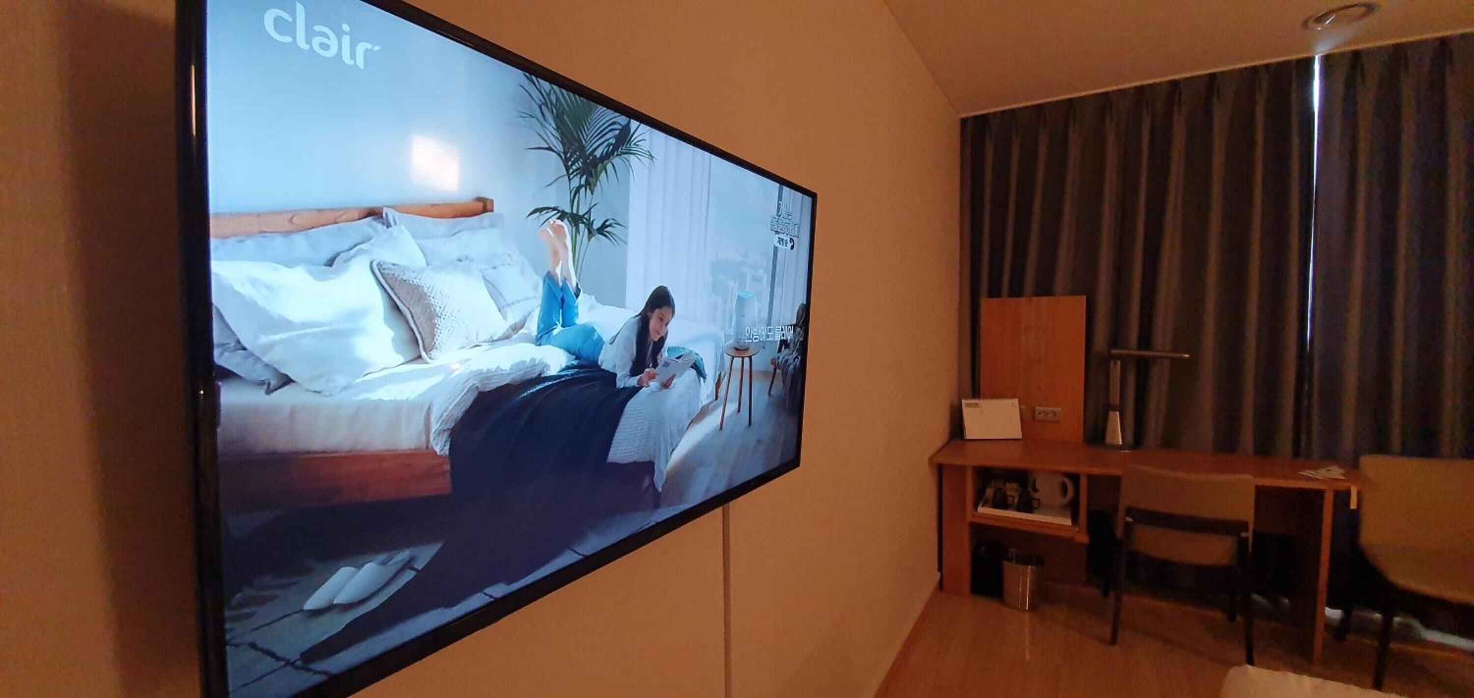 A large flat-screen TV mounted on a wall displaying a room with a bed, a plant, and a desk with a chair.